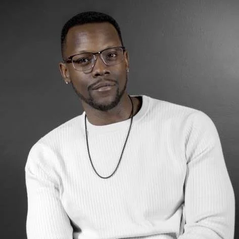 Portrait of a man with black hair, glasses, and a beard, wearing a white sweater and a necklace, against a gray background.