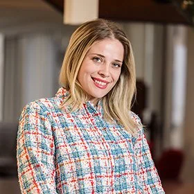 A young woman with blonde hair and a bright smile, wearing a colorful checkered blazer, standing in an indoor setting.