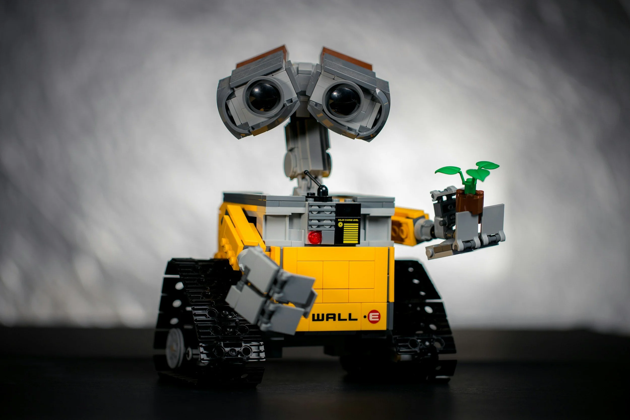 A LEGO model of WALL-E from the animated movie, holding a small plant in a gray tray. The robot has yellow and gray parts, with black treads and expressive eyes.
