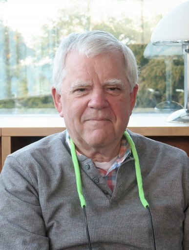 An older man with gray hair, wearing a gray shirt with a checkered collar, sitting indoors near a window with sunlight and a lamp in the background.