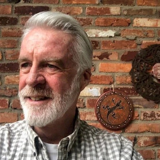 A smiling elderly man with white hair and beard stands in front of a brick wall, with a decorative clock and object hanging behind him.
