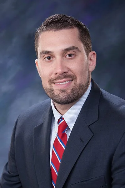 Professional portrait of a man in a dark suit, light blue shirt, and red and blue striped tie, smiling against a dark mottled background.