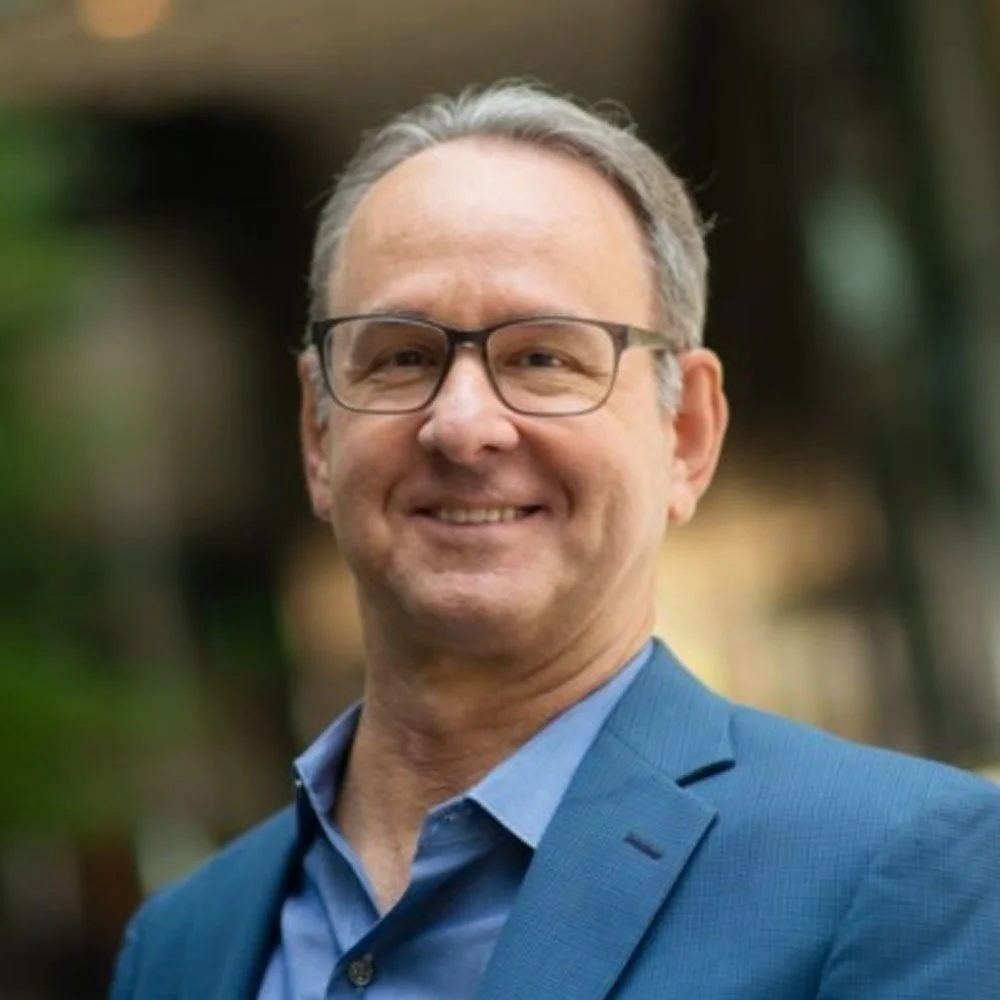 A middle-aged man with gray hair, wearing glasses and a blue suit, smiling outdoors with a blurred background.