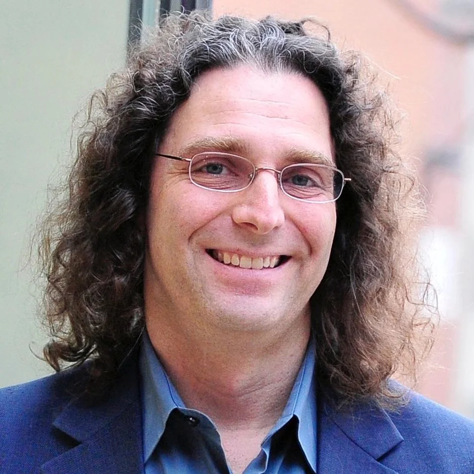A man with long, curly brown hair, glasses, and a friendly smile, wearing a dark blue blazer and a light blue shirt.