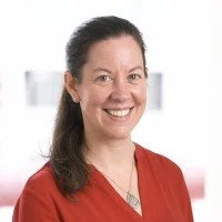 A woman with dark hair tied back, smiling, wearing a red top, in an indoor setting with blurred background.