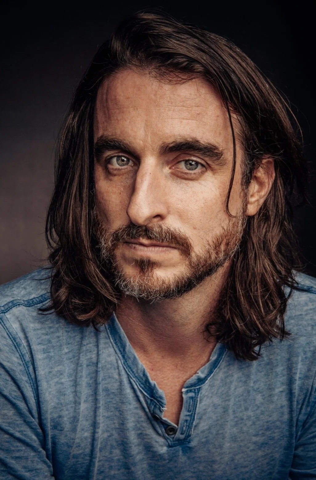 Close-up portrait of a man with long brown hair, blue eyes, and a beard, wearing a blue henley shirt against a dark background.