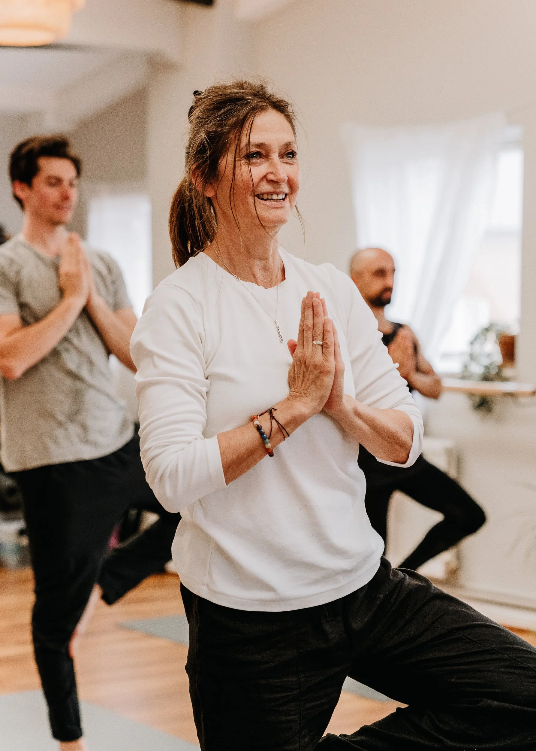 A group of people practicing yoga at Every Body Studio in Oxford, with a smiling woman in the foreground doing a yoga pose with her hands together in prayer position.