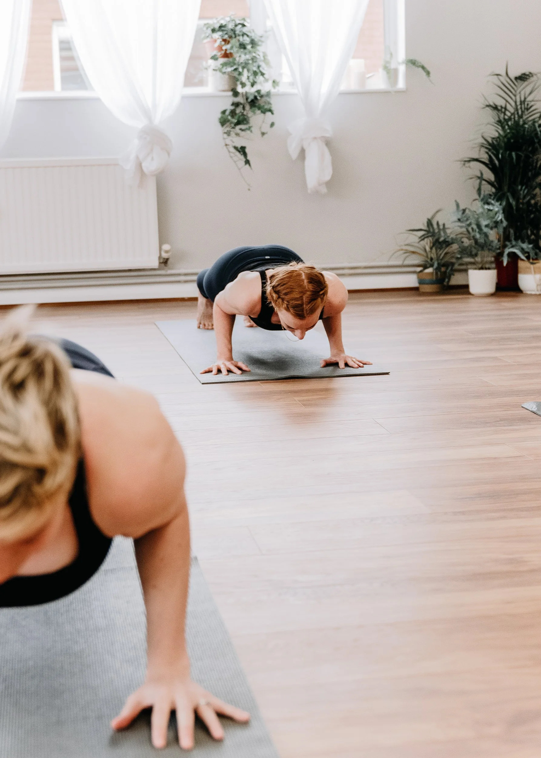 Two women practicing yoga in a bright room at Every Body Studio in Oxford with large windows, white curtains, and potted plants, in a plank pose on yoga mats.