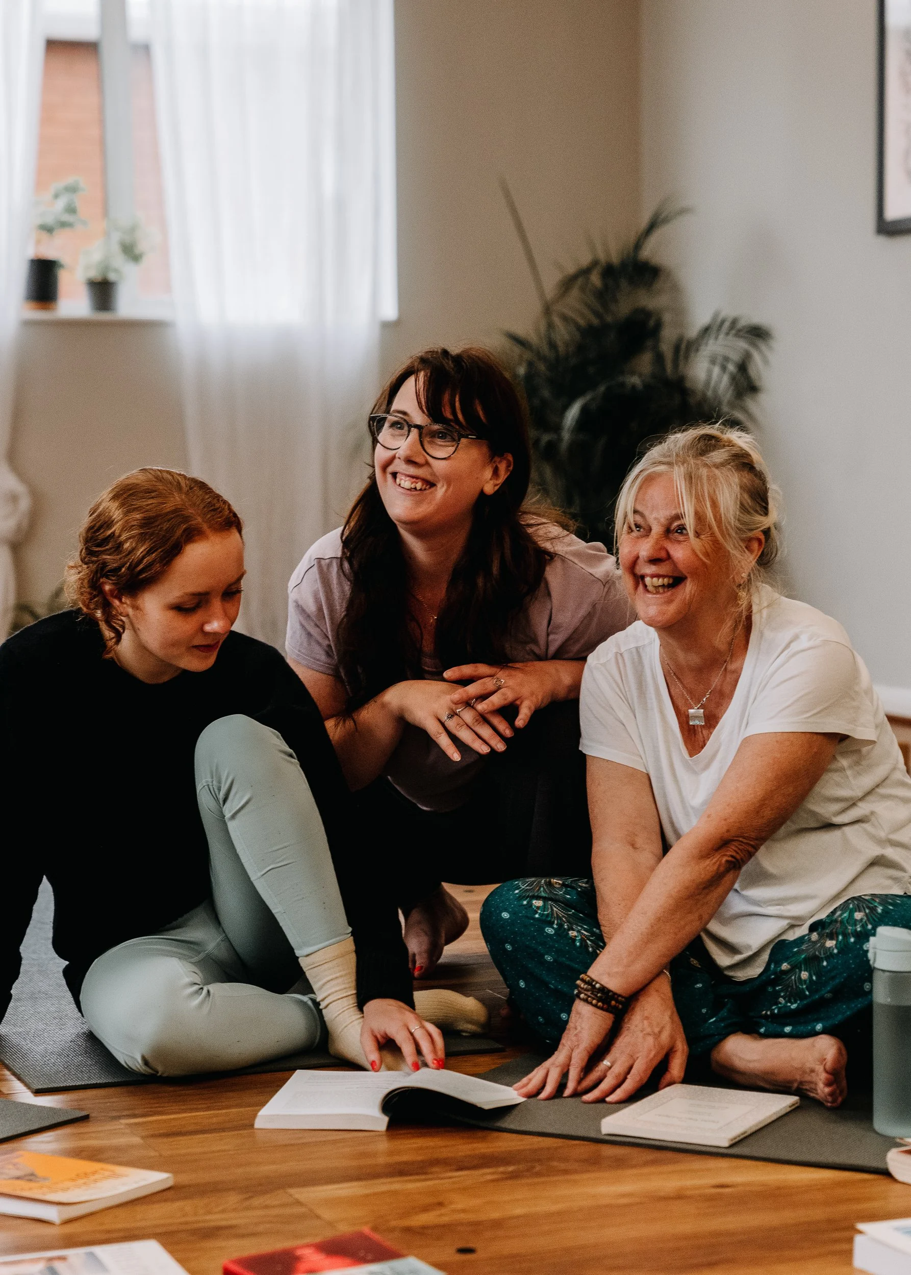Three women sitting on a wooden floor in a cozy room, sharing a joyful moment during their yoga teacher training at Every Body Studio in Oxford. One woman is reading from a book, while the other two are smiling and engaged.