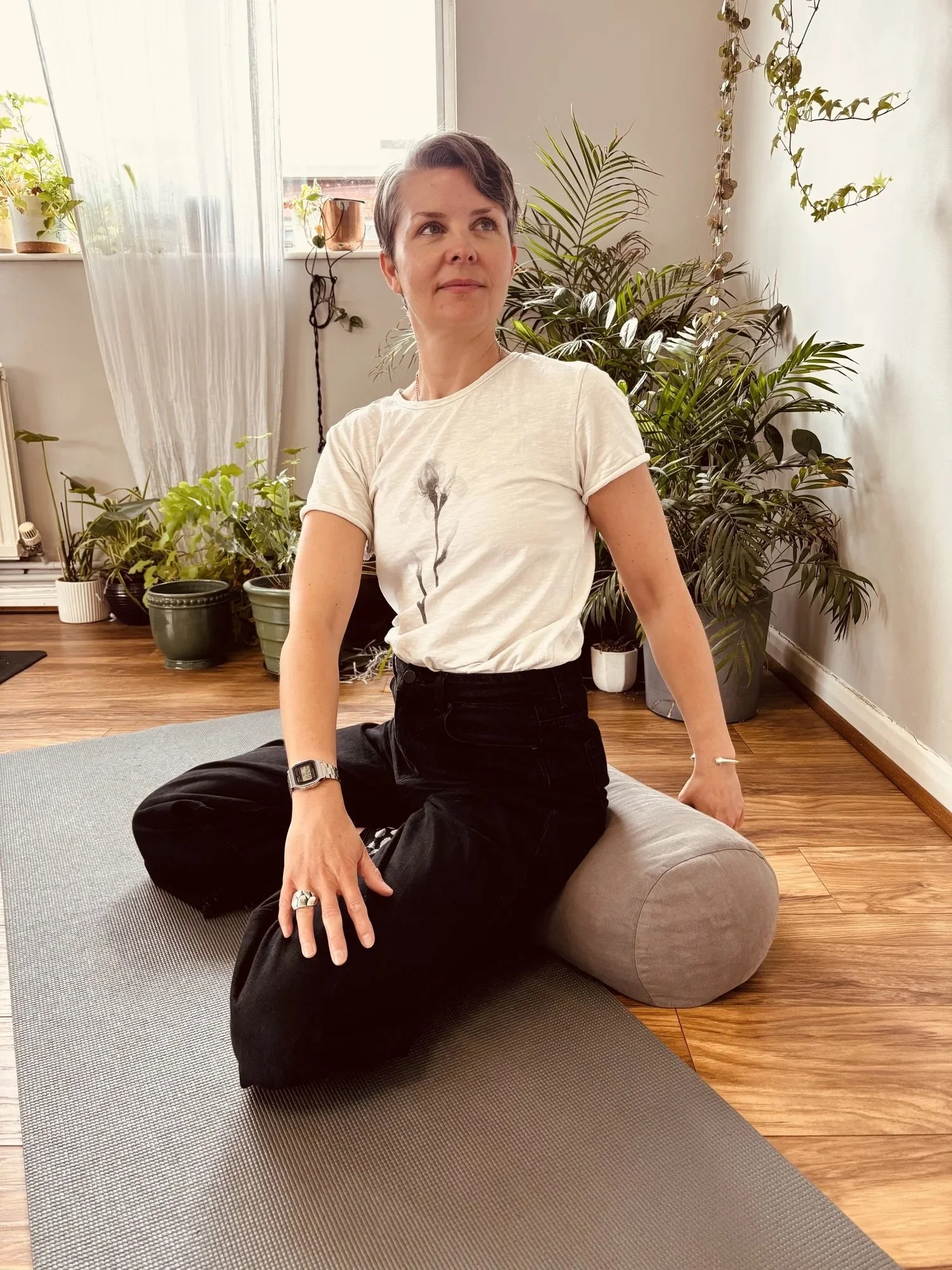 A woman with short dark hair sitting cross-legged on a yoga mat indoors, surrounded by green houseplants near a window with sheer curtains at Every Body Studio Oxford