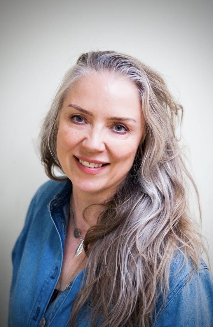 Portrait of a smiling woman with long, wavy gray-blonde hair wearing a blue denim jacket and a necklace against a plain light-colored background at Every Body Studio Oxford