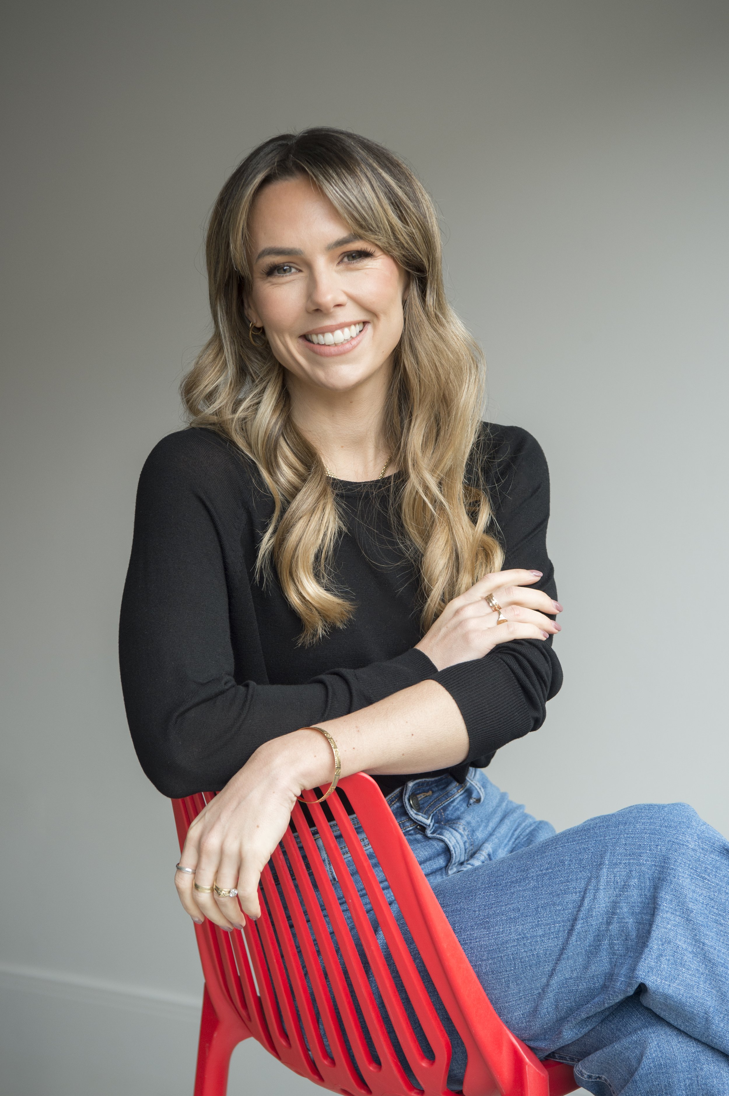 Smiling woman with long, wavy hair sits on a red chair. She wears a black top and jeans, exuding a relaxed and friendly vibe against a neutral background.