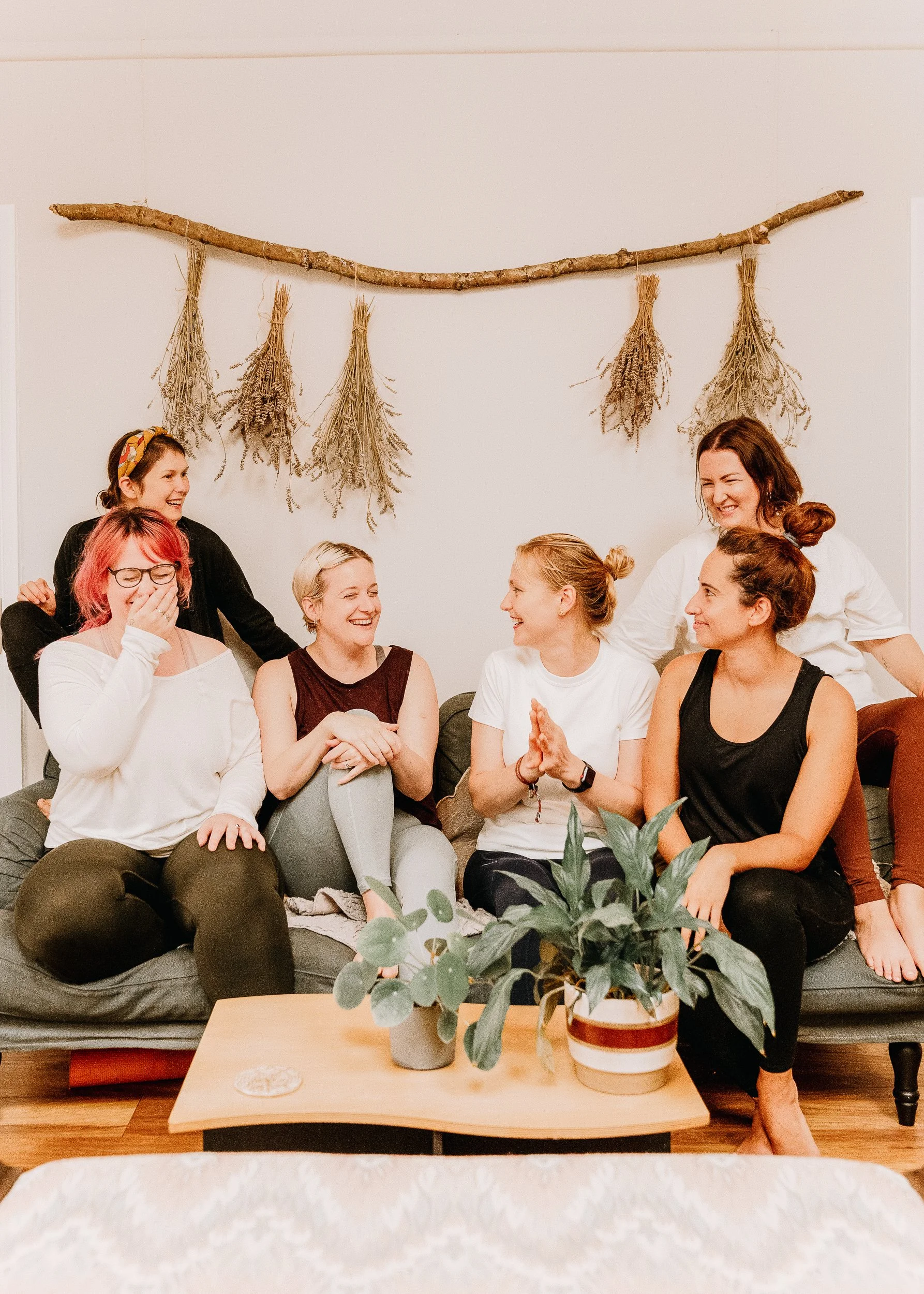 Seven women sitting on a gray couch in a cozy living room, laughing and chatting. A wooden coffee table with potted plants in front of them, and a wall hanging made of a stick and dried herbs in the background.