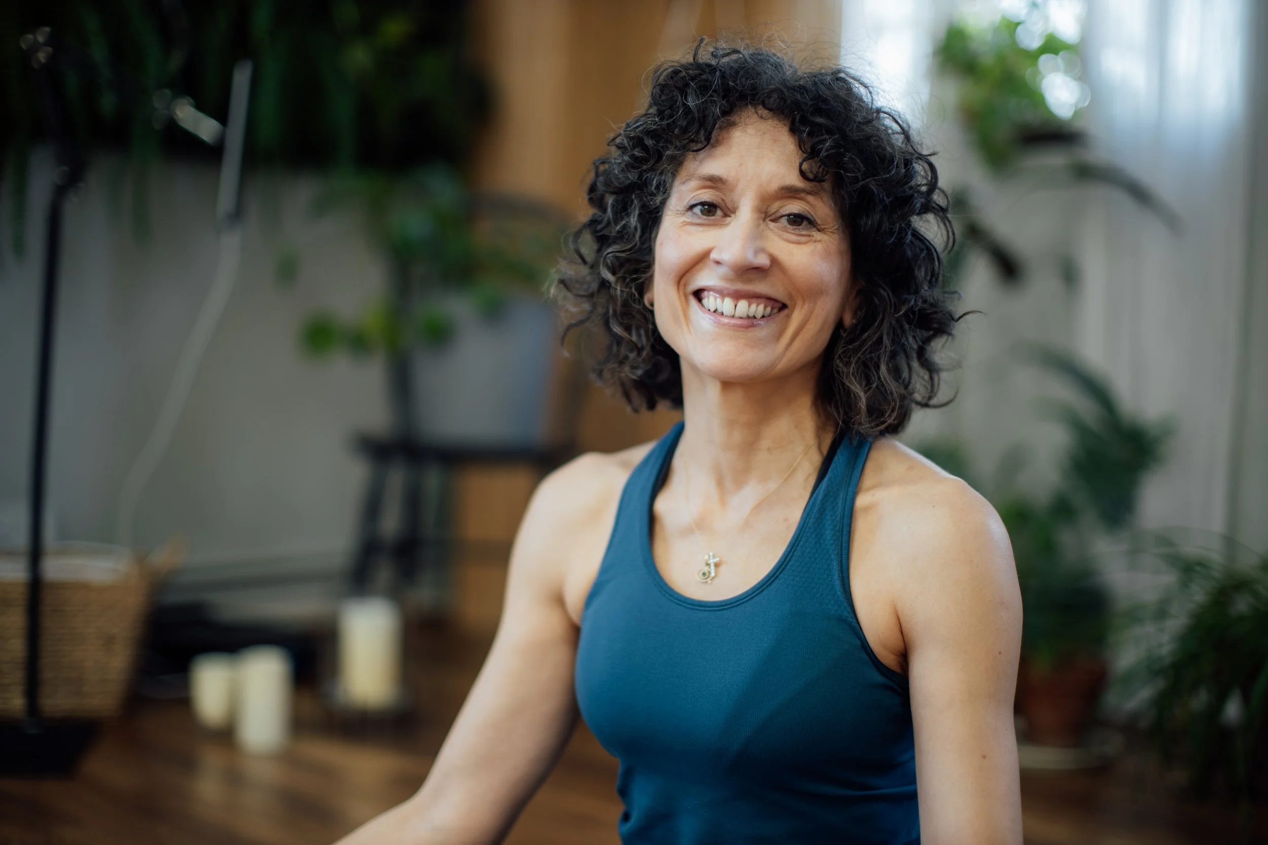 A woman with curly dark hair smiling at the camera, wearing a blue tank top and a necklace with a cross and a circle, indoors with plants and candles in the background at Every Body Studio Oxford