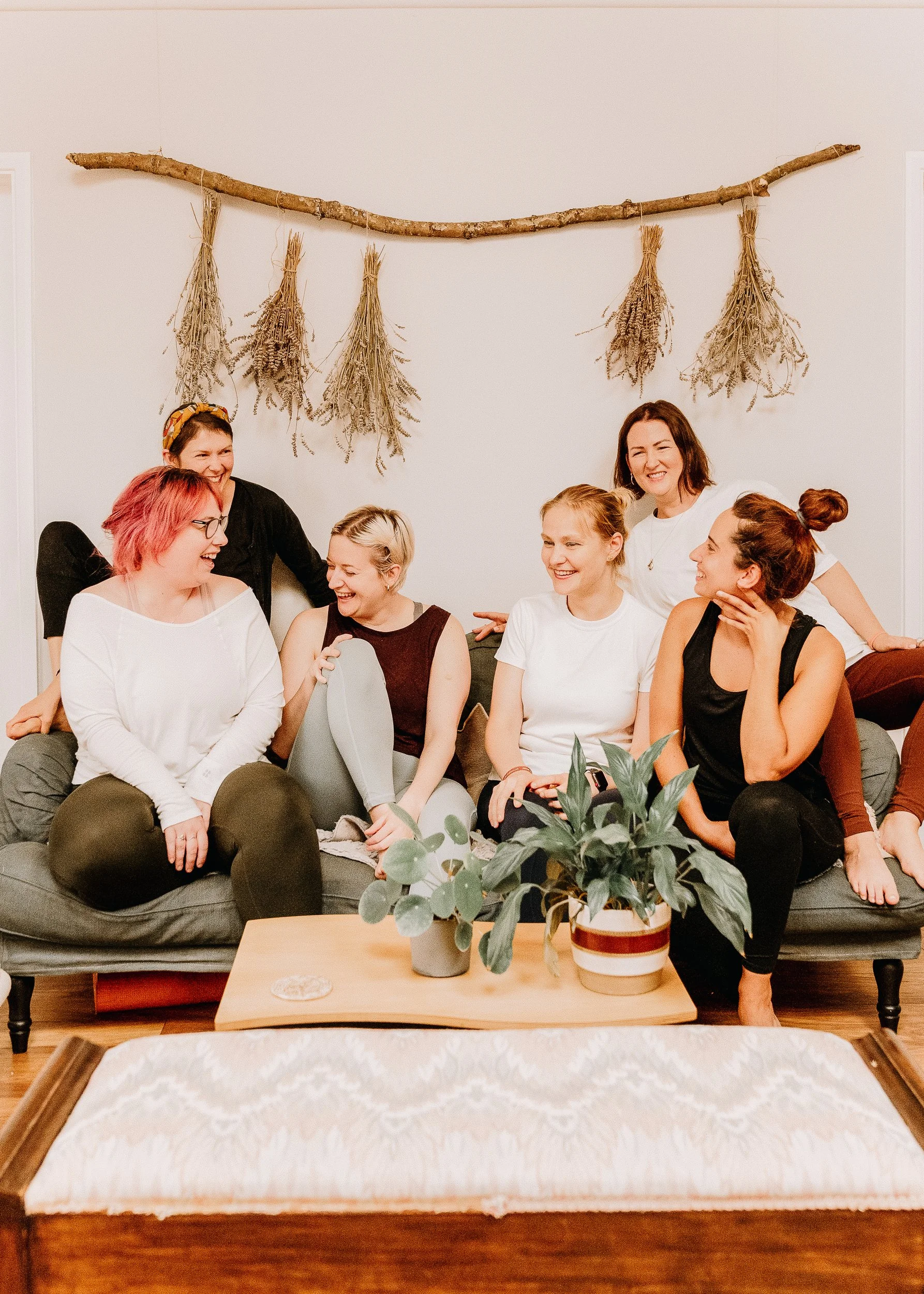 Six women sitting and standing on a couch, laughing and talking in a cozy living room with a white wall, dried herbs hanging on a stick above them, and potted plants on a wooden coffee table.