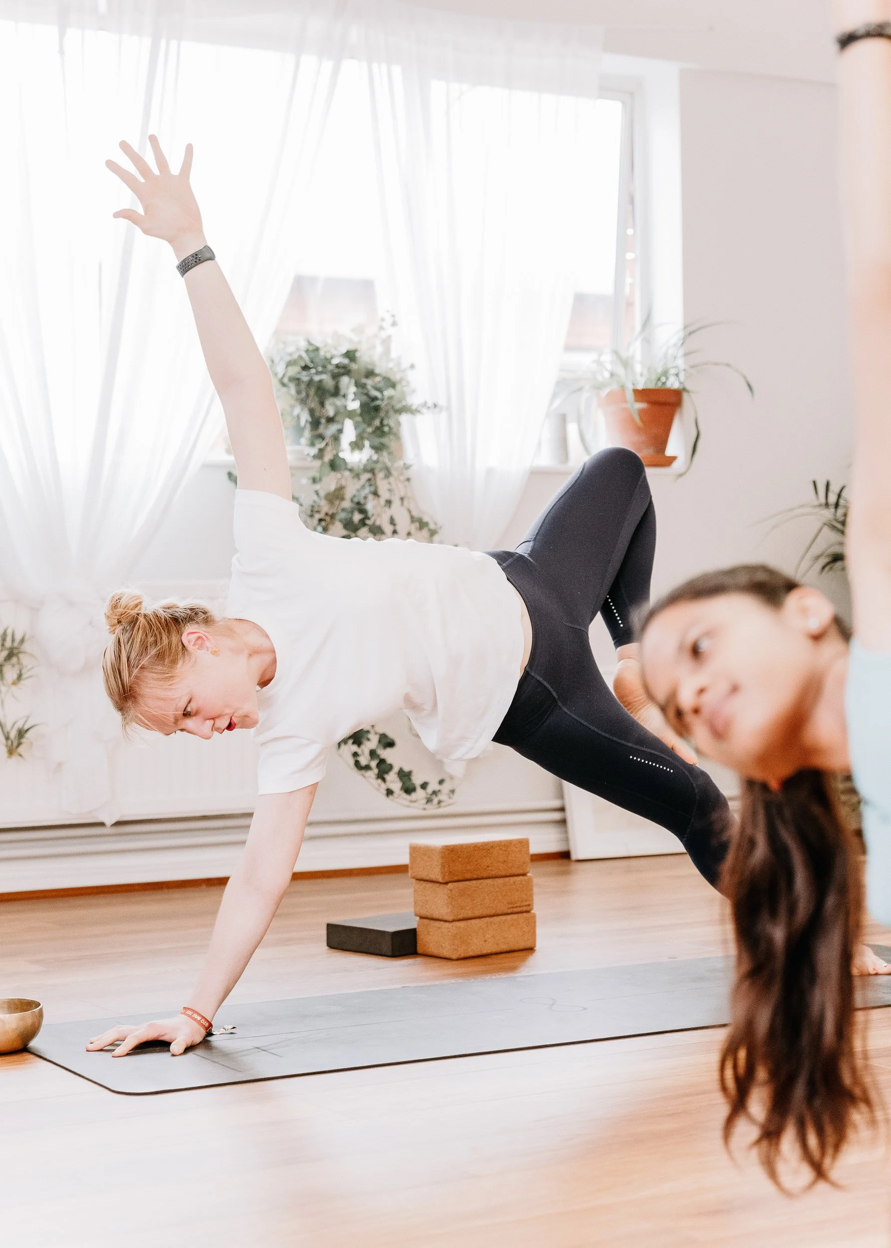 A woman practicing yoga in a bright room at Every Body Studio in Oxford, supporting herself on a yoga mat with one hand and one foot, while extending the opposite arm and leg. She is wearing a white t-shirt and black leggings.