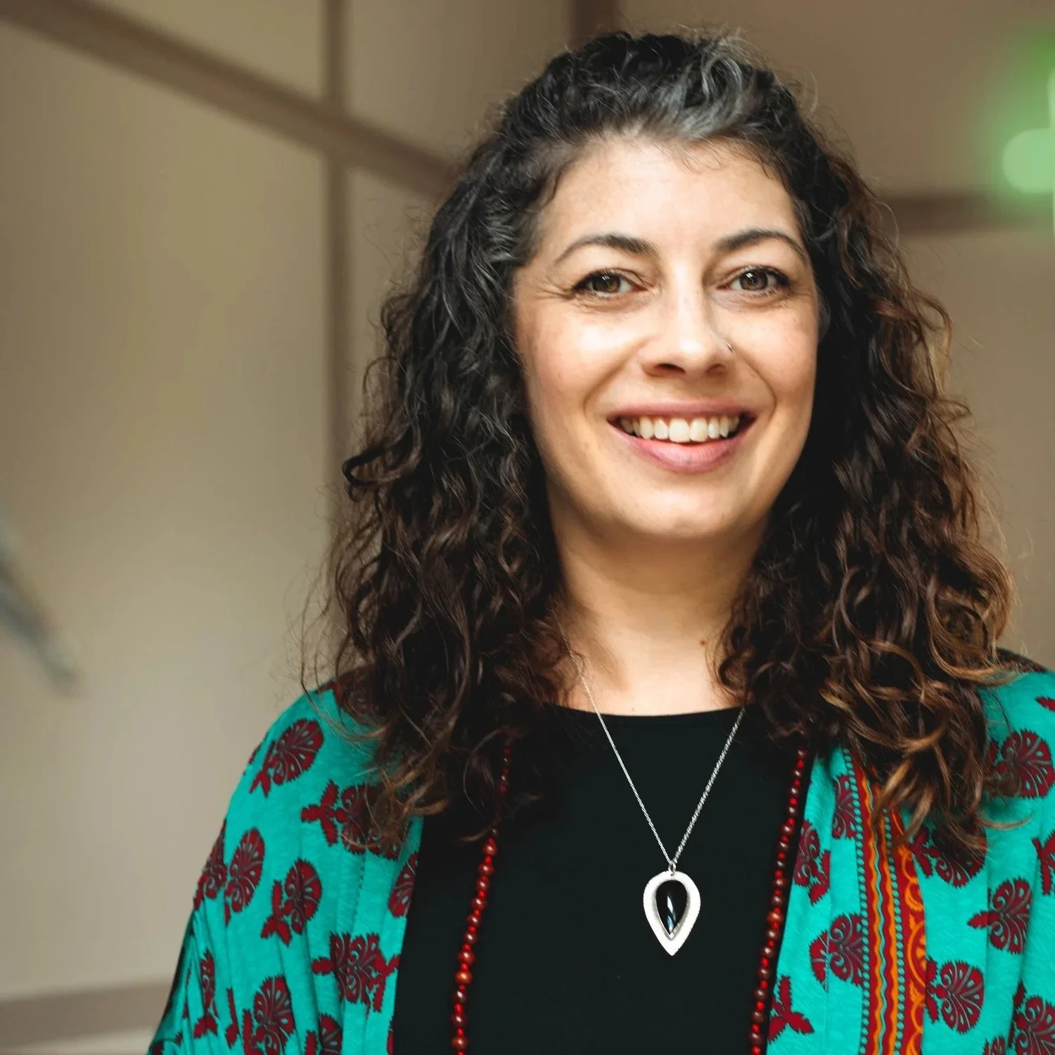 A woman with curly dark hair smiling, wearing a black top, a teal patterned shawl, a silver necklace with a heart-shaped pendant, and a red beaded necklace indoors.