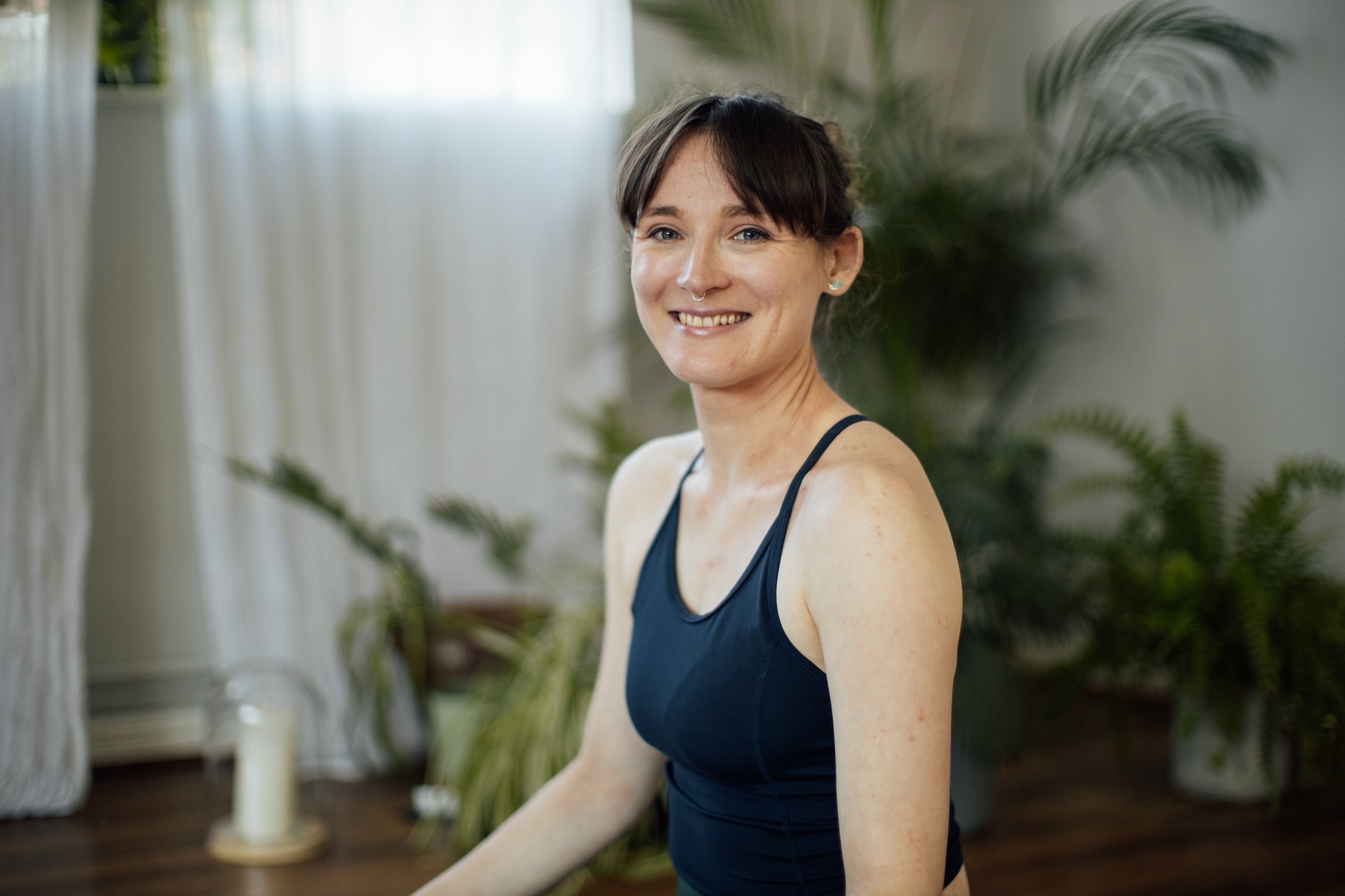 A smiling woman in a black tank top sitting indoors with green plants and white curtains in the background at Every Body Studio Oxford