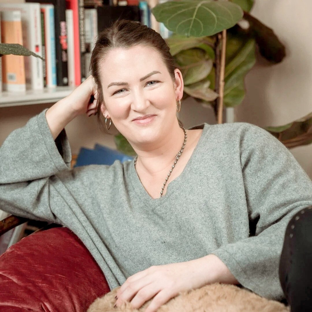 A woman with dark hair pulled back, wearing a gray sweater and jewelry, sitting on a red cushion, smiling at the camera with a bookshelf and large leafy plant in the background at Every Body Studio Oxford