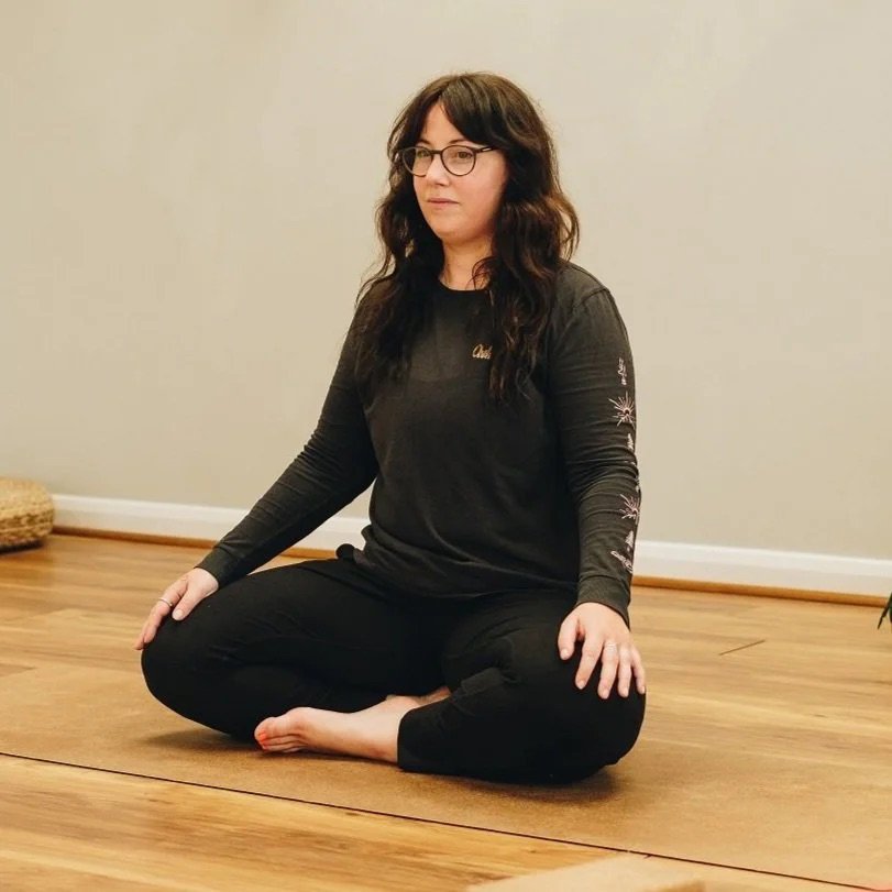 Woman with glasses and dark hair sitting cross-legged on a wooden floor in a meditation pose in front of a plain wall at Every Body Studio Oxford