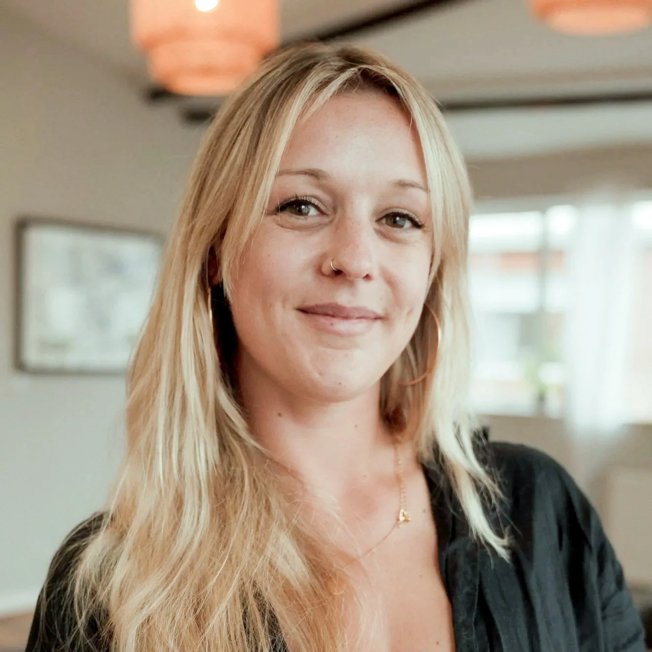 A young woman with long blonde hair, wearing a black top and a gold necklace, smiling in a warmly lit room at Every Body Studio Oxford
