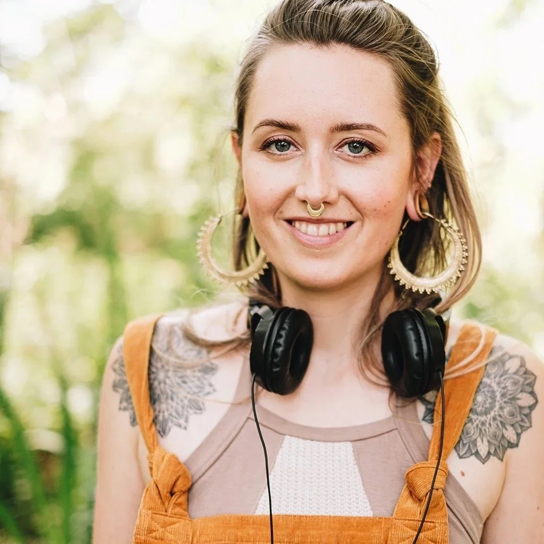 A young woman with tattoos on her shoulders, wearing large hoop earrings, a septum piercing, and headphones around her neck, smiling outdoors with blurred green foliage in the background.