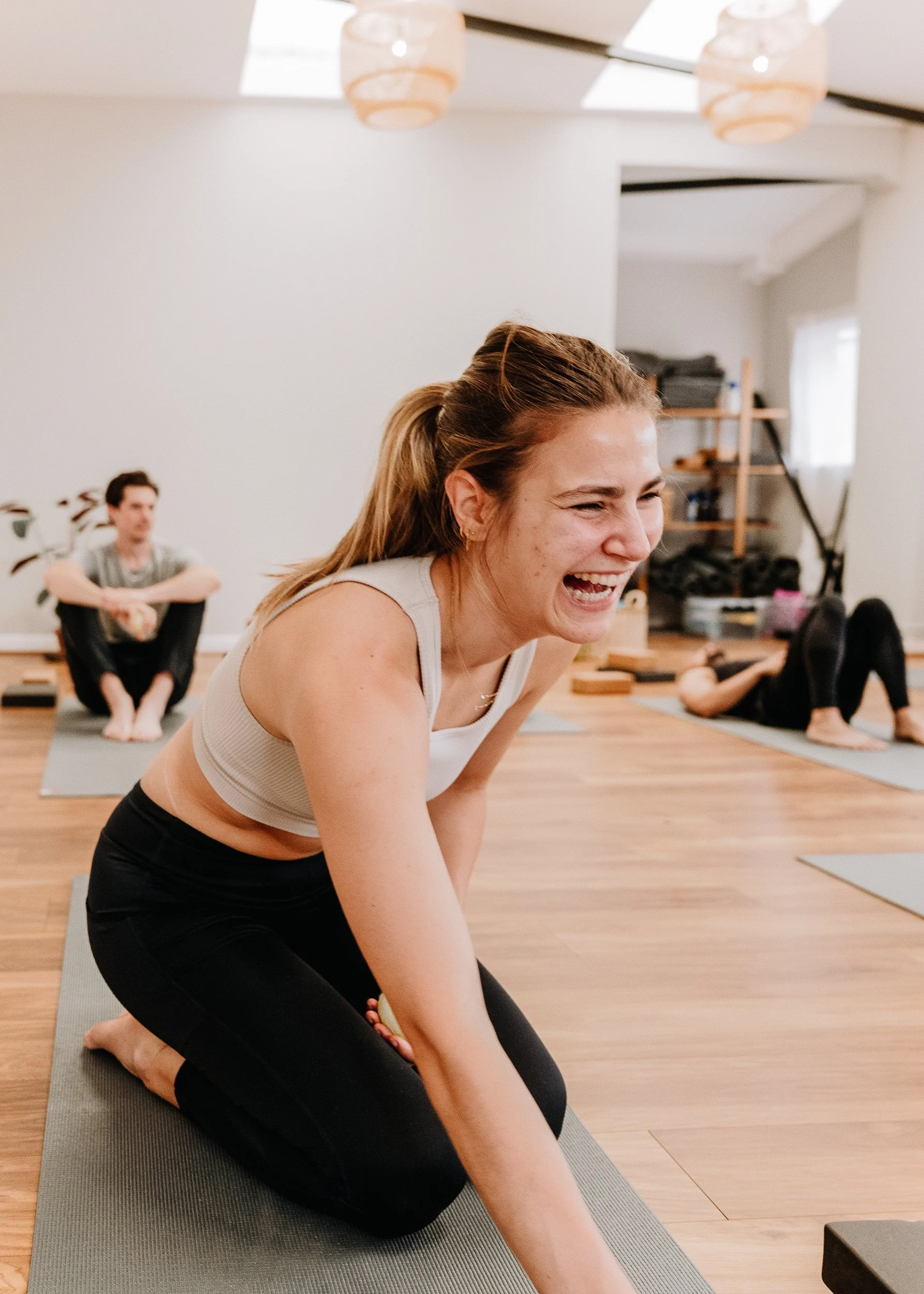 A woman participating in a yoga class at Every Body Studio in Oxford, smiling and laughing, with two other individuals in the background on yoga mats in a well-lit room.