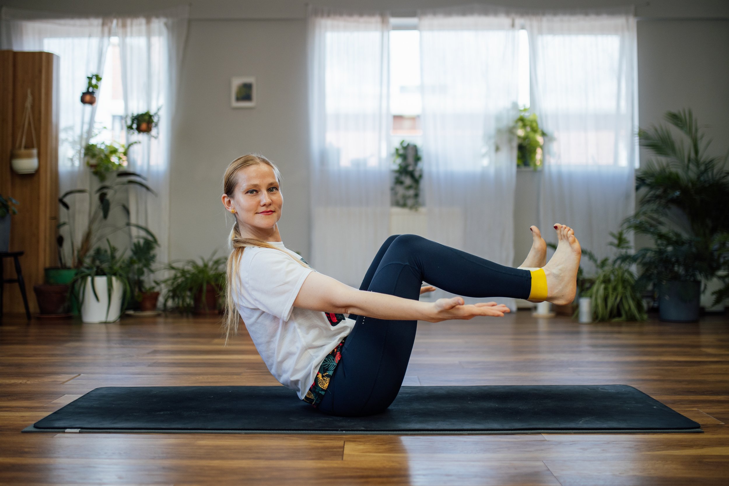 A woman practicing yoga indoors on a black mat, sitting in a boat pose with her arms extended forward and legs raised. The room has wooden flooring, plants, and sheer curtains at Every Body Studio Oxford