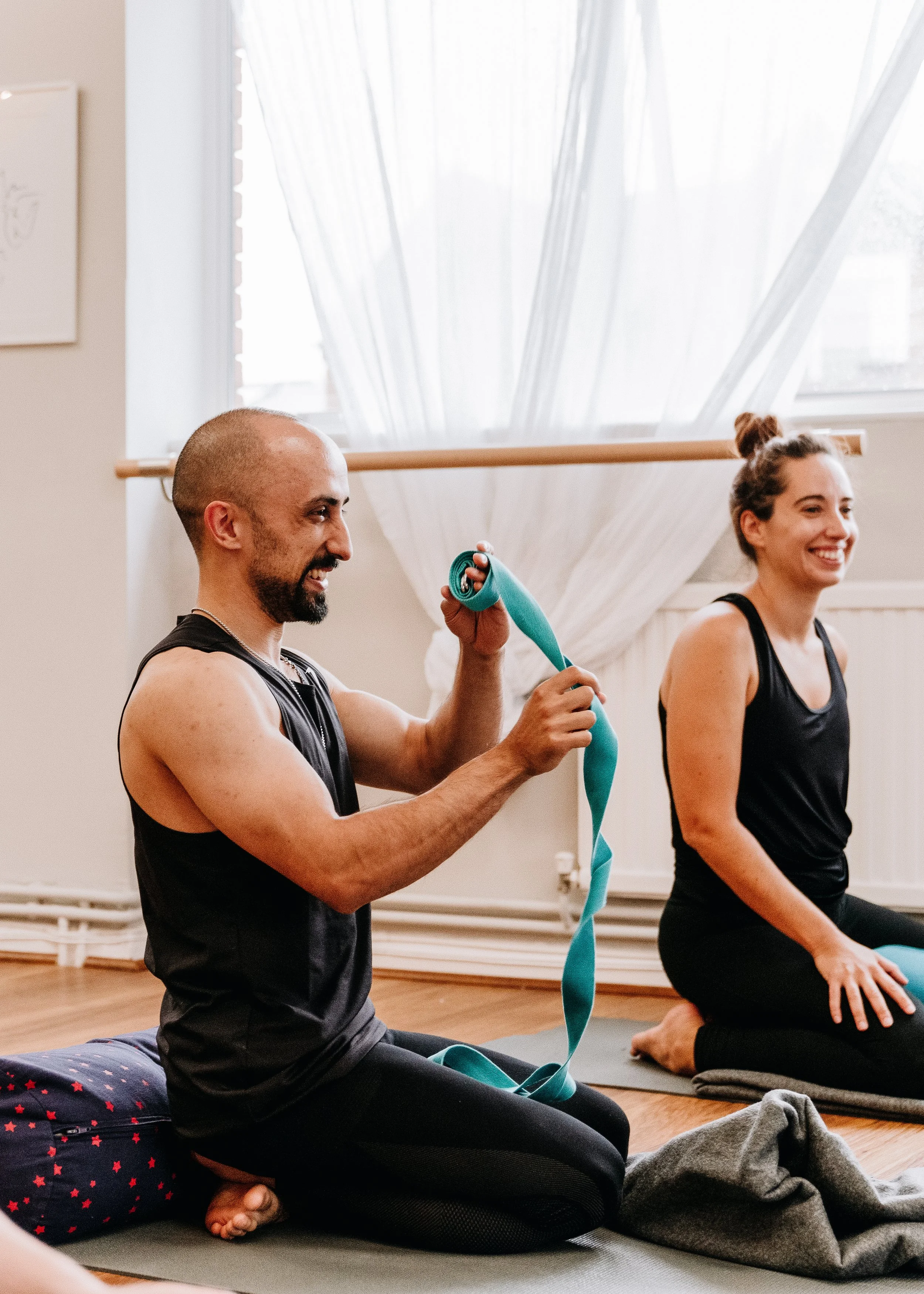 Two people practicing yoga at Every Body Studio in Oxford, kneeling on yoga mats, one wrapping a blue yoga strap, with a window and sheer curtains in the background.