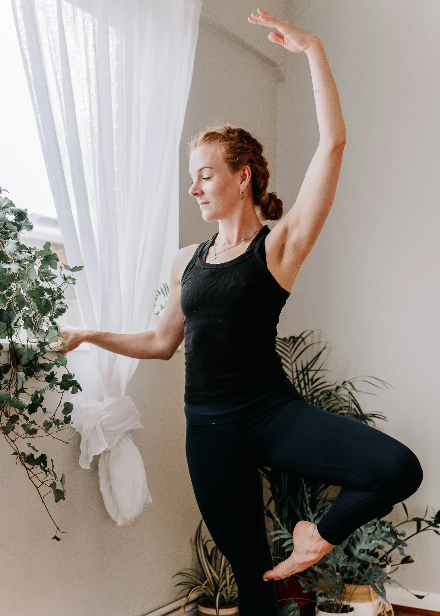 A woman practicing barre at Every Body Studio in Oxford, wearing a black tank top and black leggings, balancing on one foot with her eyes closed and arm raised.