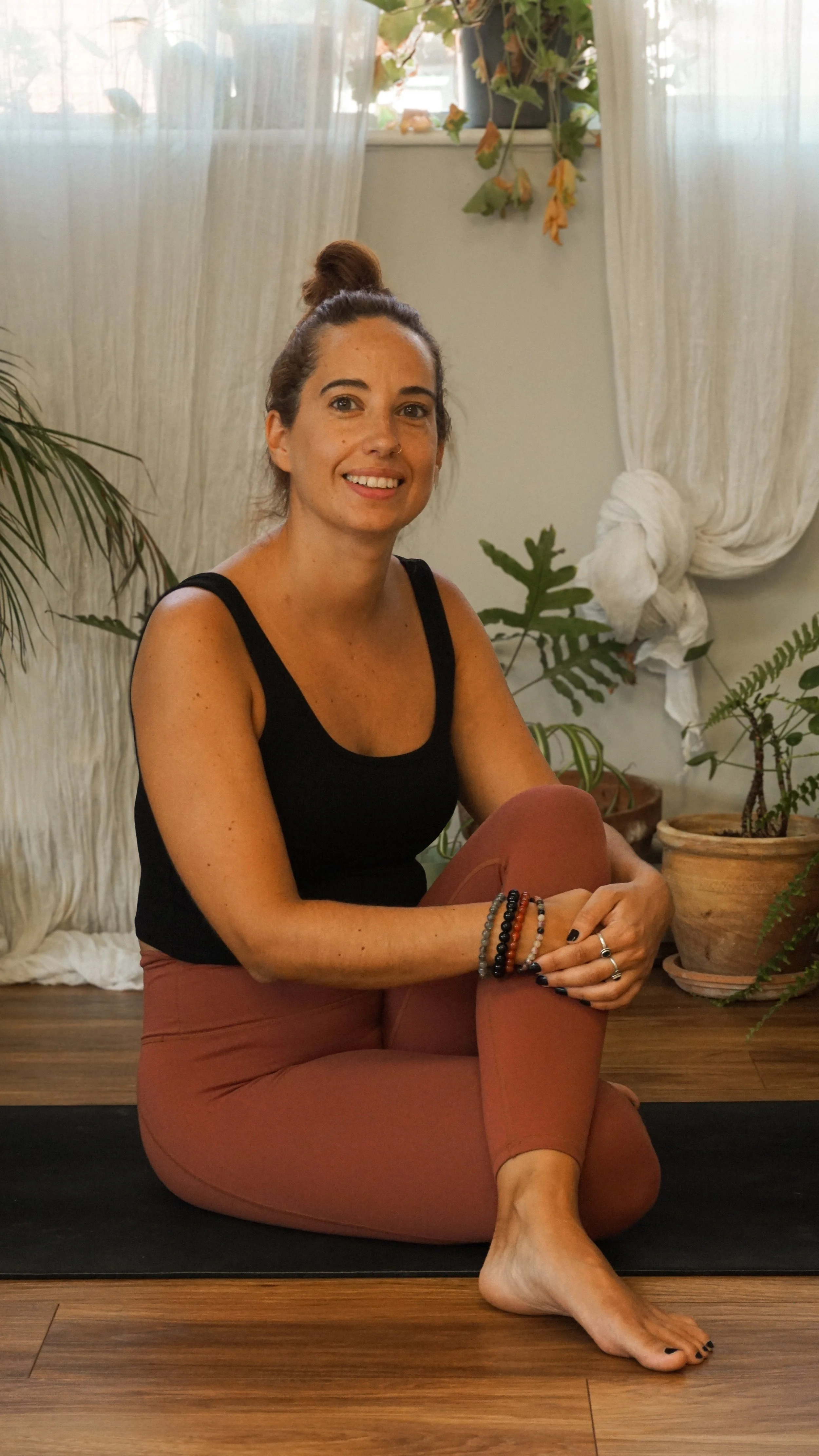 A woman practicing yoga indoors, sitting on a yoga mat with her legs crossed, smiling at the camera. She is wearing  black tank top and mauve leggings, with a bun in her hair, surrounded by indoor plants and white curtains at Every Body Studio Oxford