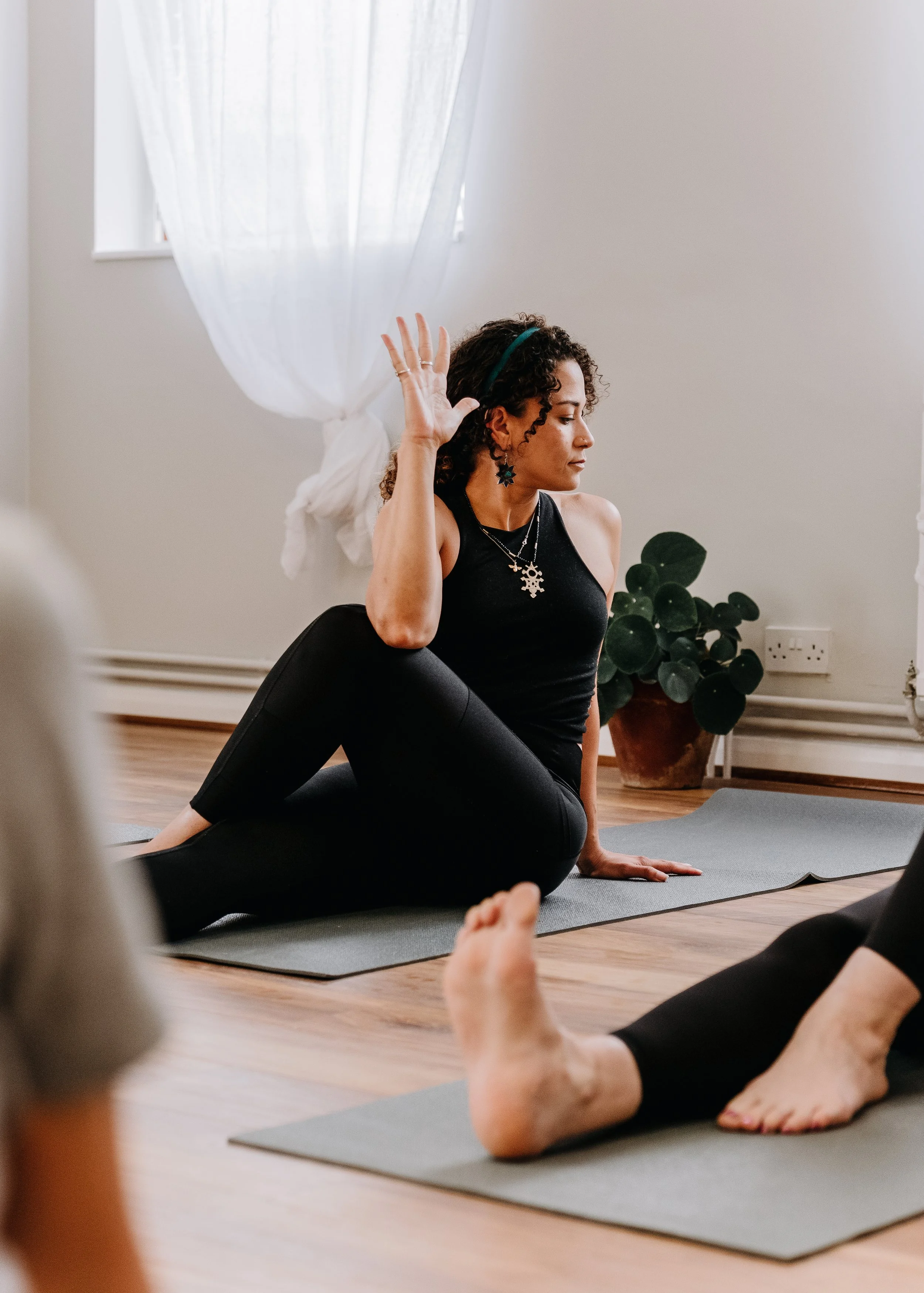 A woman leading a yoga class at Every Body Studio in Oxford, seated on a yoga mat in a bright room with natural light, white curtains, and a potted plant, demonstrating a seated twist pose with her hand raised.
