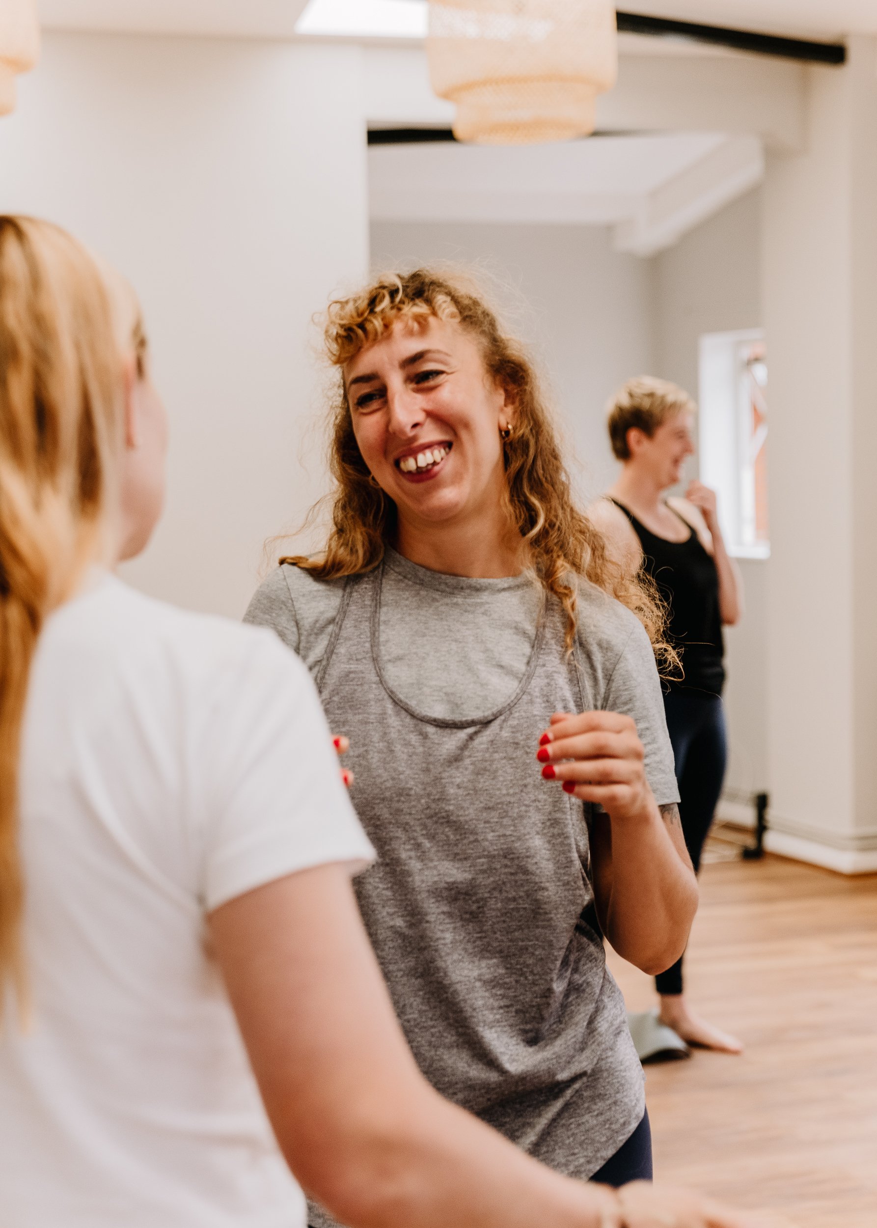 Three women are in a well-lit room, engaging in conversation. The woman in the foreground has curly blonde hair, a gray t-shirt, and is smiling while speaking. In the background, another woman with short hair and a black tank top is smiling and appears to be listening. The third woman, partially visible, has long hair and is wearing a white shirt. The room has light-colored walls, wooden flooring, and hanging ceiling lights.
