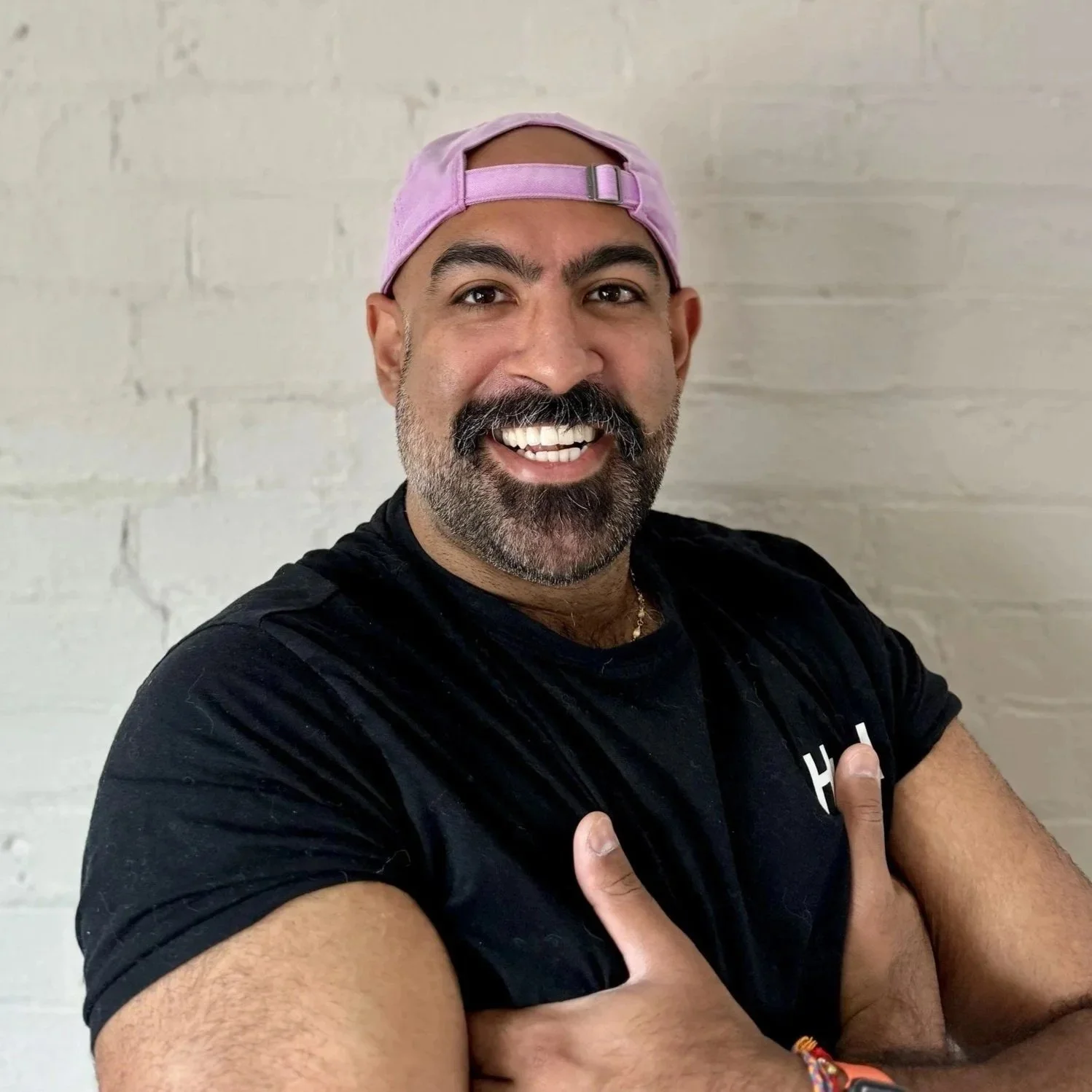 A man with a beard and mustache wearing a pink baseball cap backward, black T-shirt, and colorful bracelet, smiling and crossing his arms in front of a white brick wallat Every Body Studio Oxford