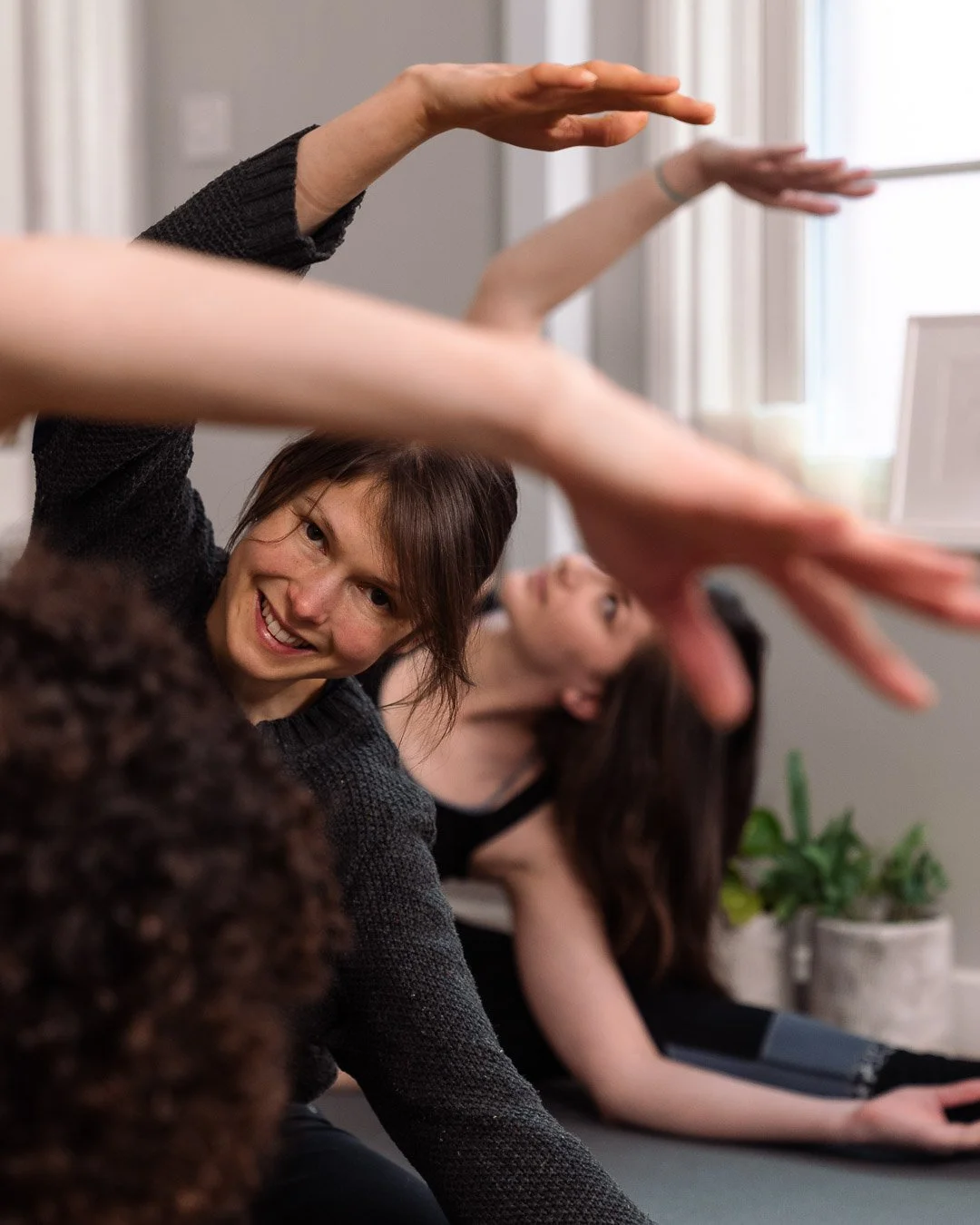 Three women practicing yoga indoors, reaching forward with their arms extended, with one smiling woman in the foreground at Every Body Studio Oxford