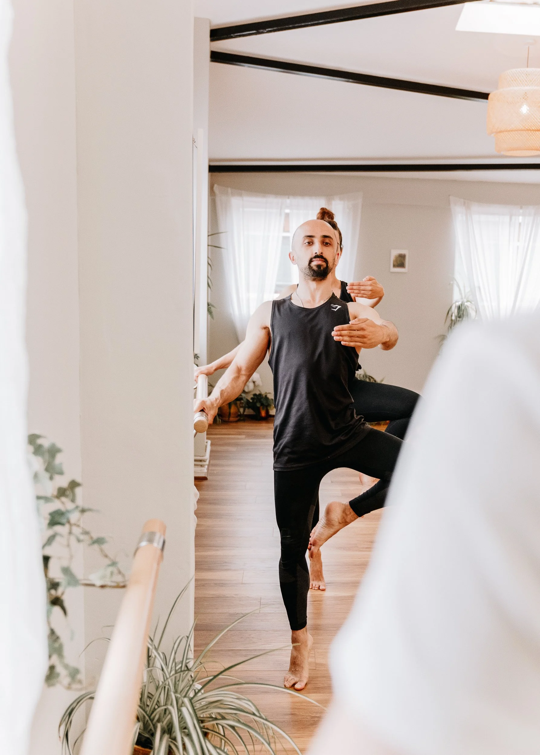 A man practicing yoga at Every Body Studio, holding a yoga pose near a mirror, with his arms extended and one leg raised, wearing a black sleeveless athletic shirt and black leggings.