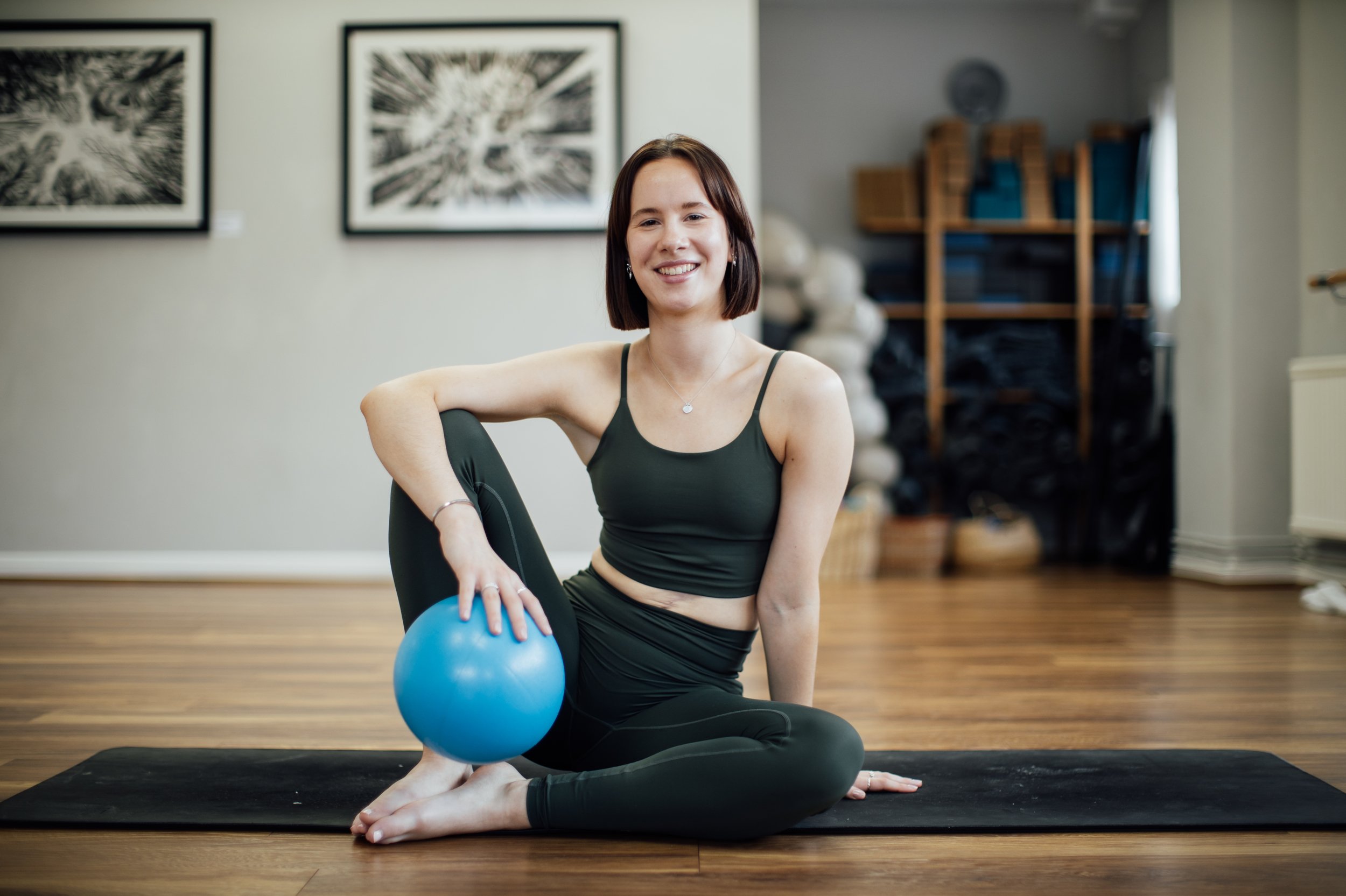 A woman with short dark hair practicing yoga indoors on a black mat, holding a blue exercise ball against her knee, smiling at the camera, with framed artwork on the wall behind her at Every Body Studio Oxford