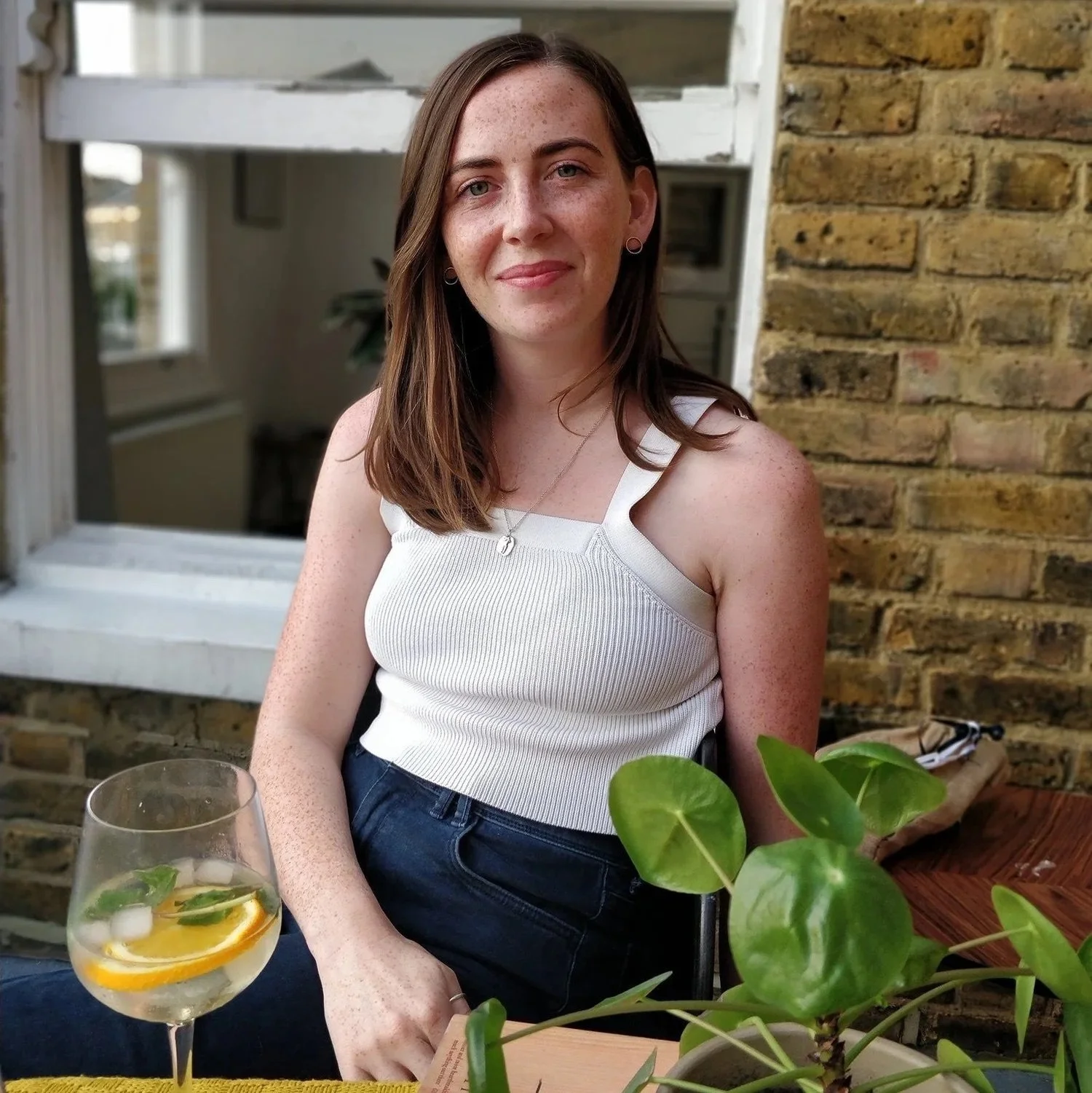A woman with shoulder-length brown hair and fair skin sitting at an outdoor table with a glass of lemonade garnished with lemon slices and mint. She is wearing a white sleeveless top and jewelry, smiling slightly, with a brick wall and window behind her.