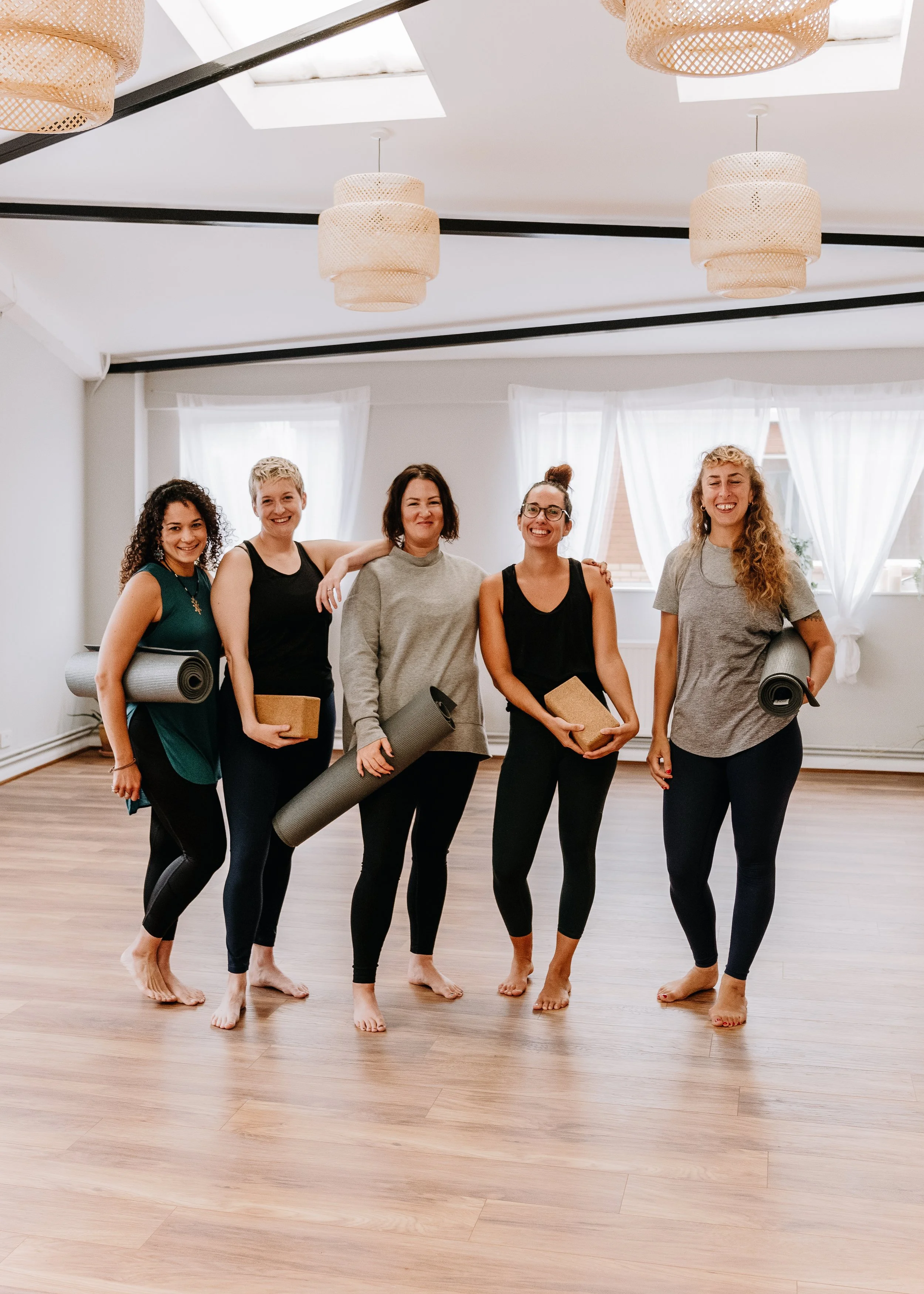 Group of five women standing barefoot in a fitness studio, holding yoga mats and blocks, smiling at the camera.