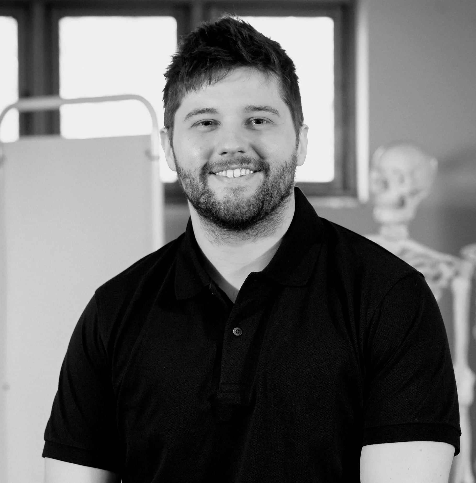 A young man with dark hair and a beard smiling, wearing a black polo shirt, with a background of windows and a skeleton model.