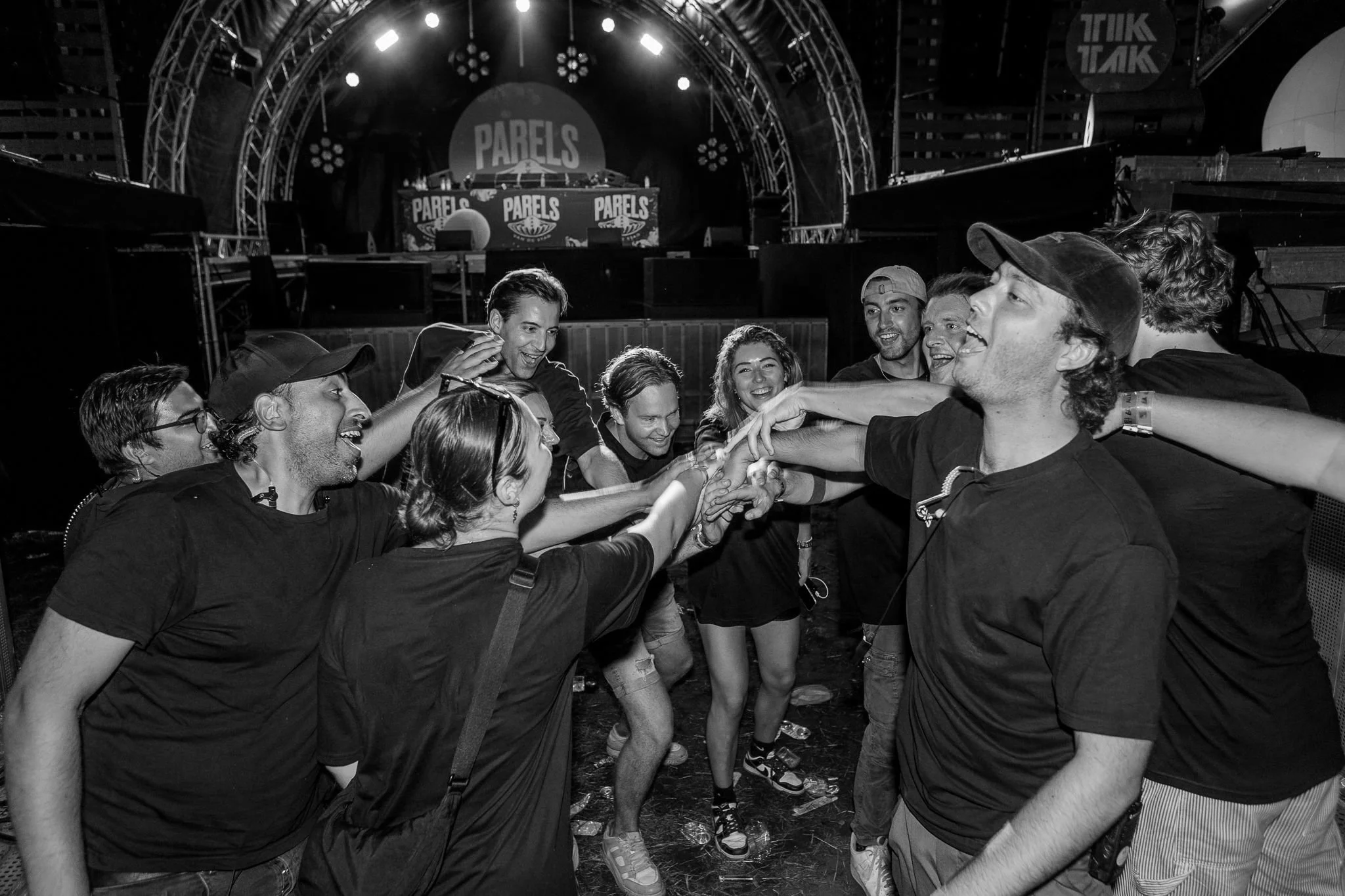 Group of people celebrating and smiling with their hands joined in a circle at an outdoor concert or festival stage.