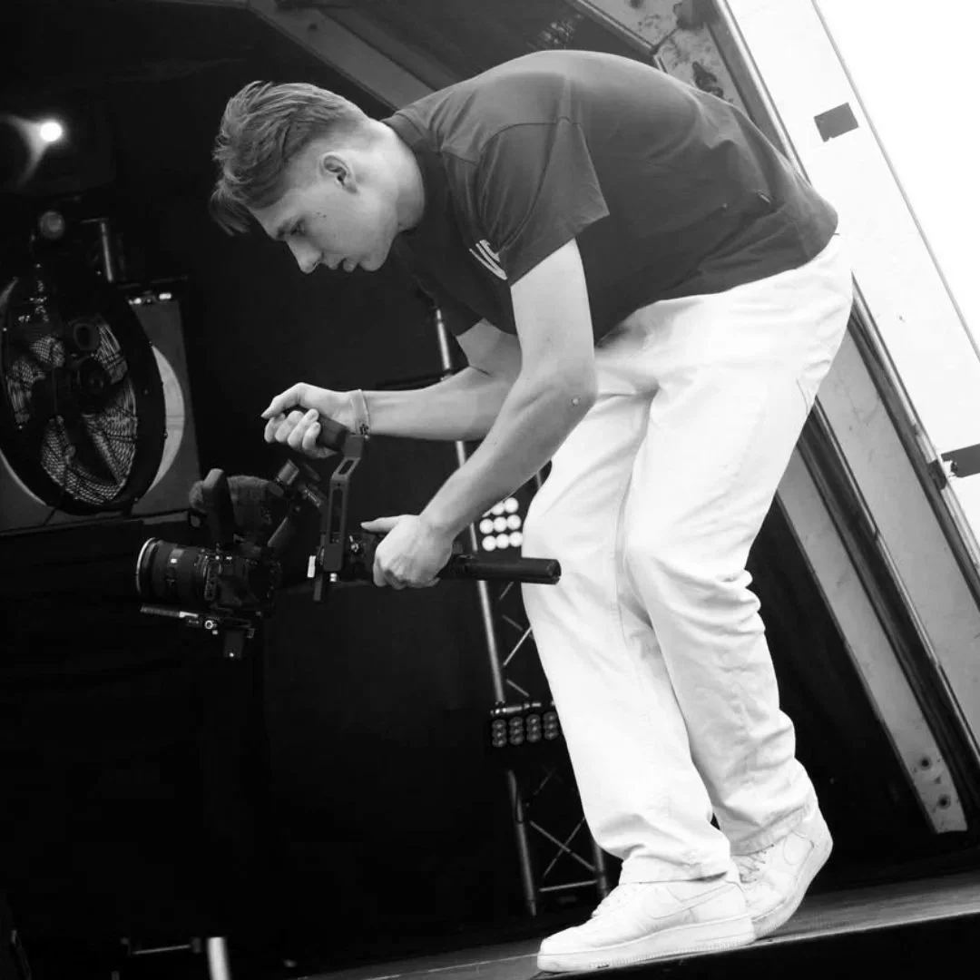 A young man operating a professional camera on a stabilizer in an indoor setting with stage lights and equipment visible.