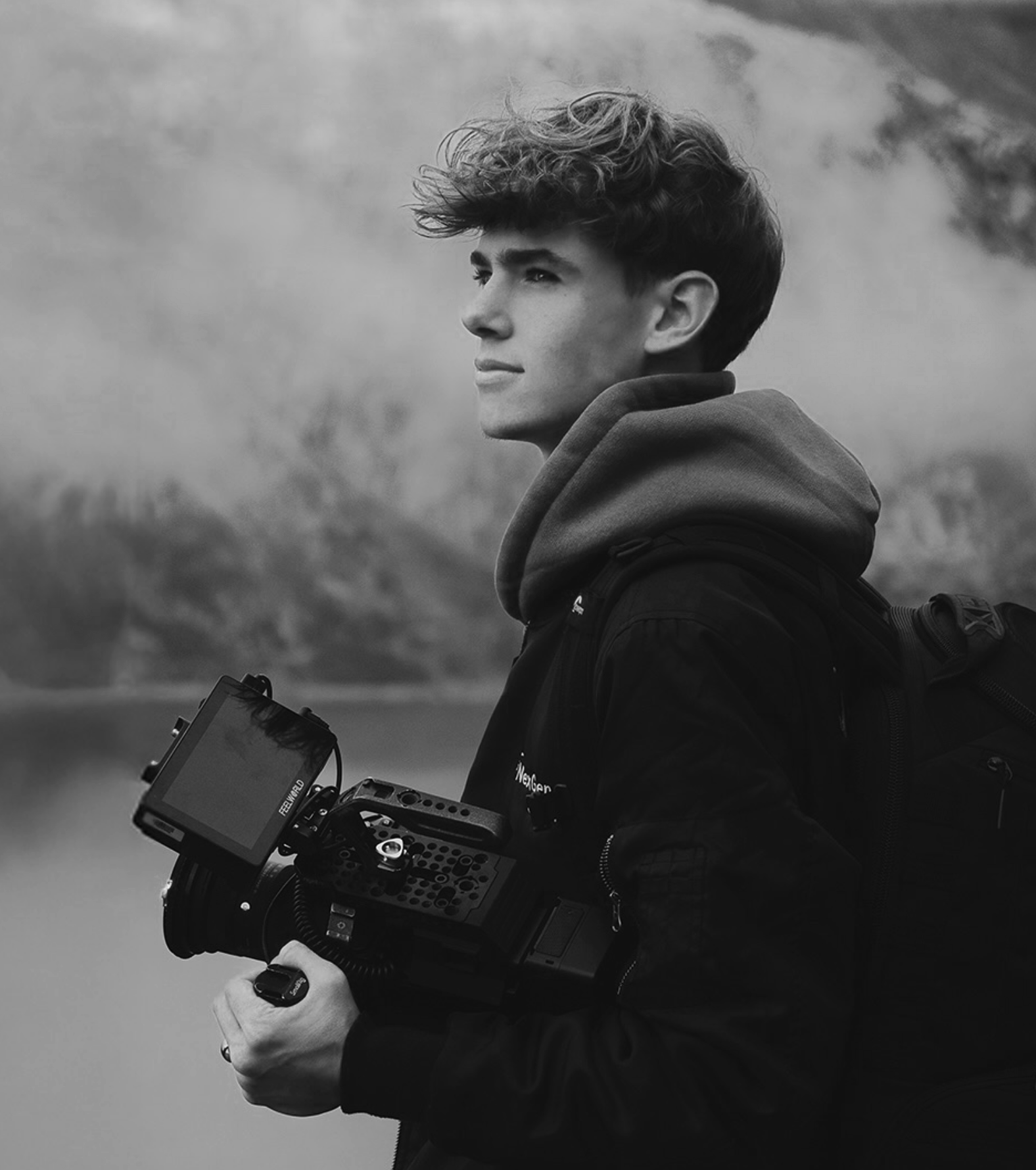 A young man with curly hair holding a professional camera, wearing a hoodie and a jacket, standing outdoors with a foggy landscape in the background.