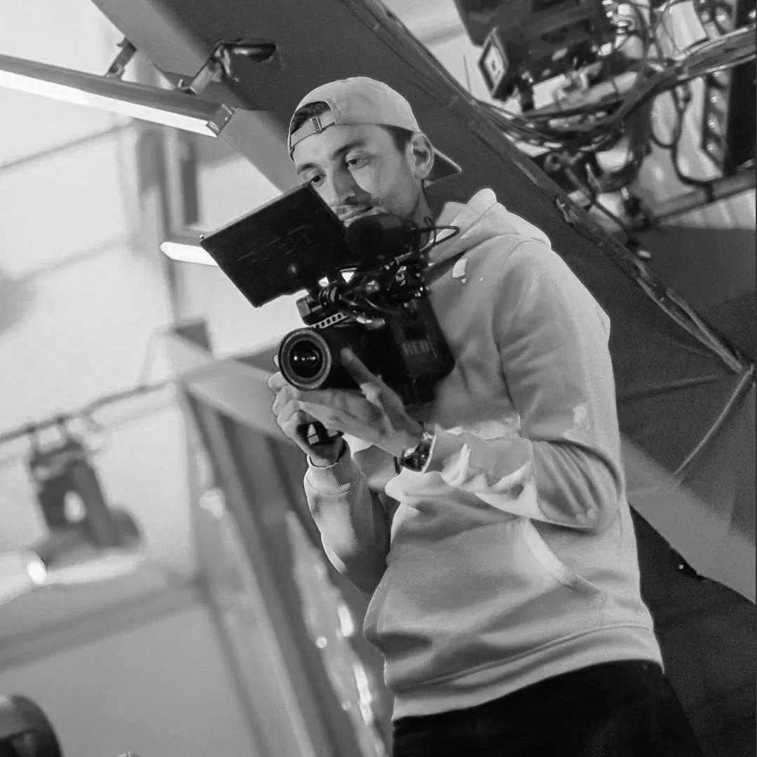 A young man wearing a backwards baseball cap and hoodie, taking a photo or video with a professional video camera.