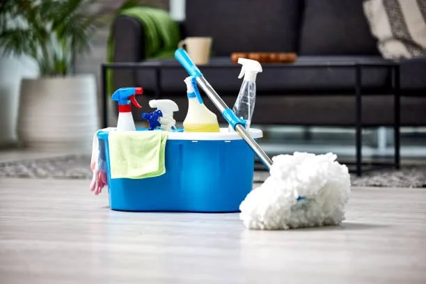 A blue plastic cleaning caddy containing spray bottles, cloths, and a mop, placed on a light-colored floor with a living room in the background.