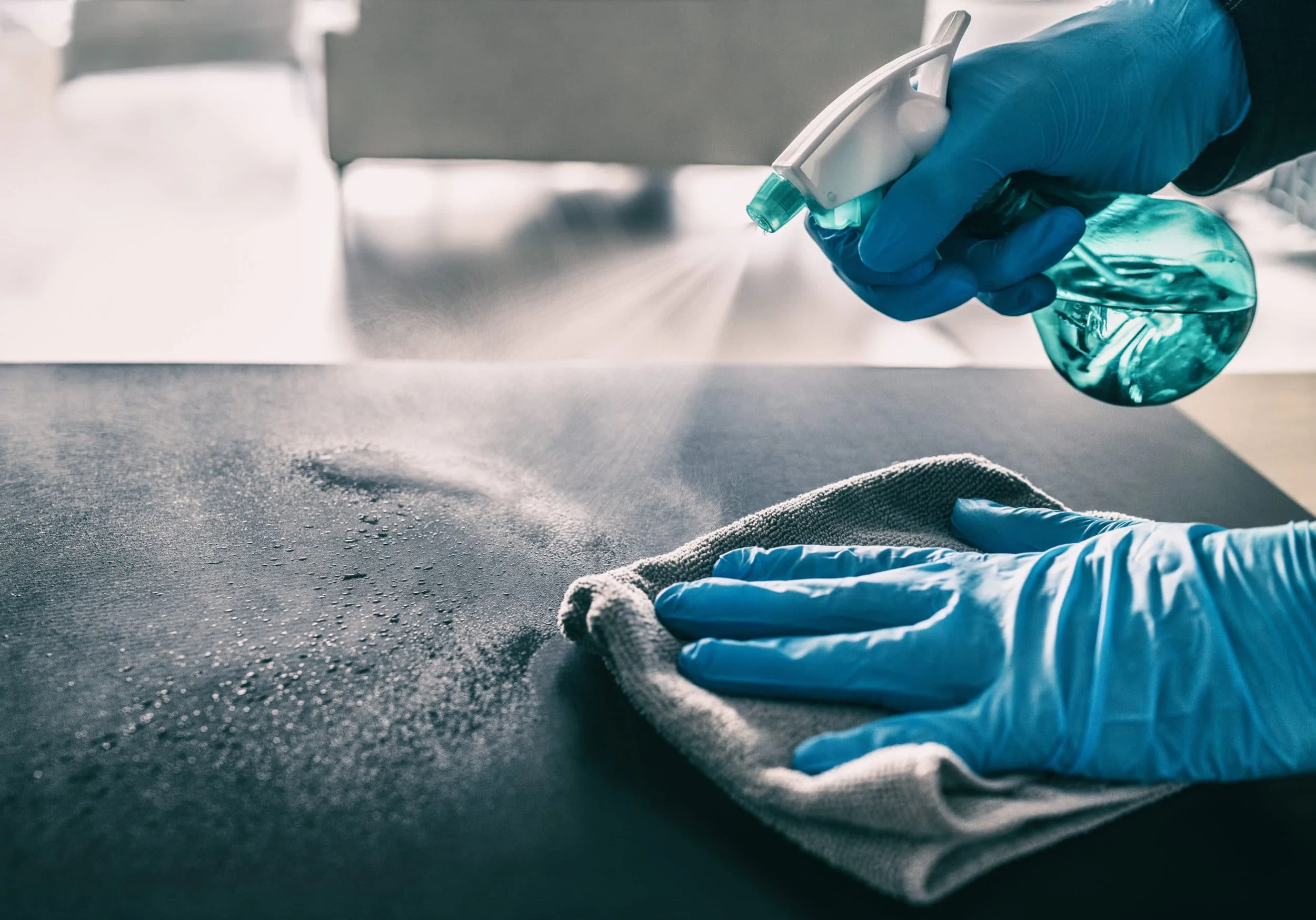 A person wearing blue gloves is spraying disinfectant onto a surface with a spray bottle, while wiping the surface with a cloth.