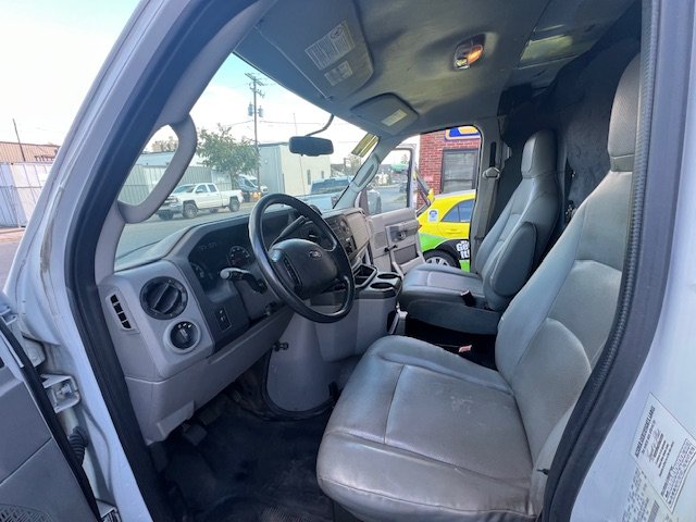 Interior of a commercial truck with two gray seats, a steering wheel, and a dashboard with various controls. Outside the window, a street scene with parked cars and buildings is visible.