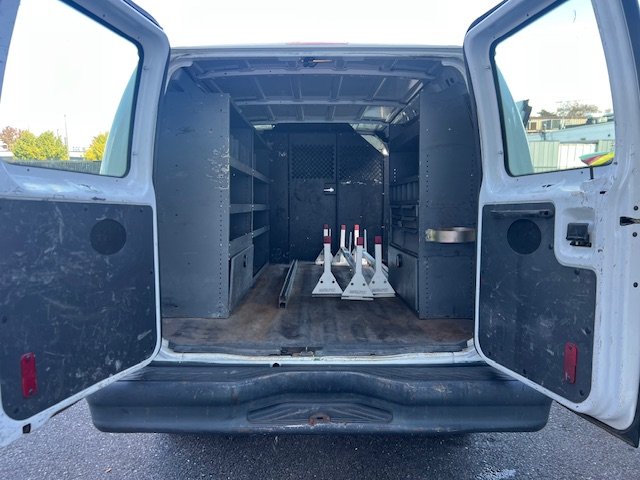 The back of a white cargo van with open doors showing an empty interior with shelves on the left and right sides, and traffic cones in the center.