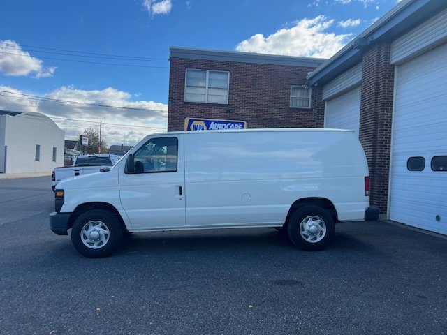 A white cargo van parked in front of an auto care shop with a brick building and blue sky in the background.