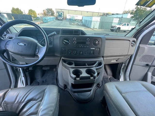 Interior of a commercial truck cab showing the dashboard, steering wheel, and front seats with view through the windshield of a yard and parked vehicles.